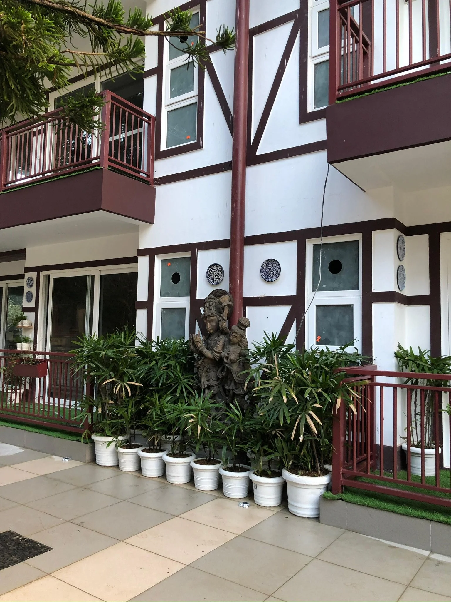 Building with red railings, white walls, and decorative brown timber framing, with potted plants and sculptures outside.