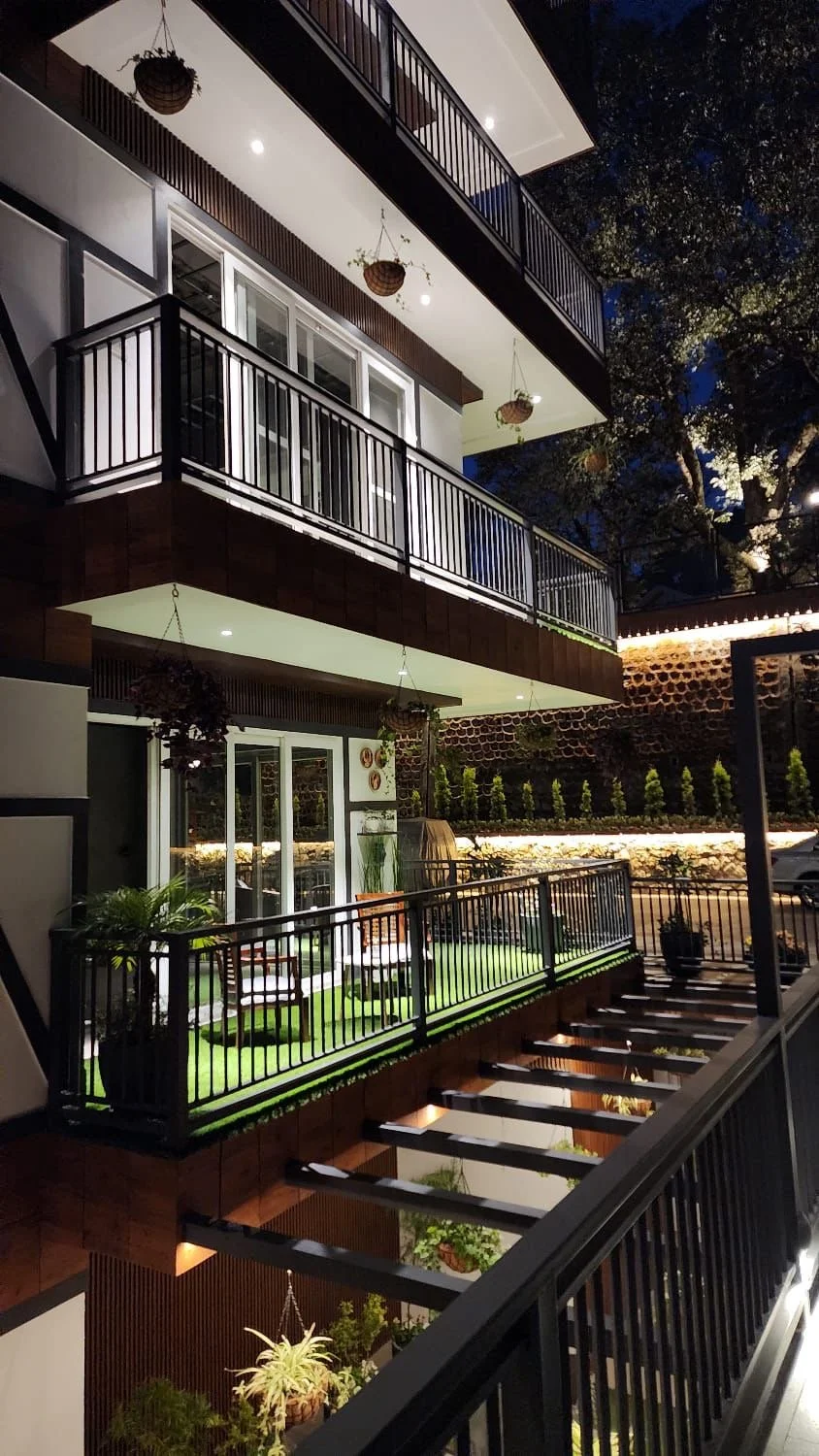Night view of a multi-story residential building with decorated balconies, hanging flower baskets, and outdoor seating area with plants.