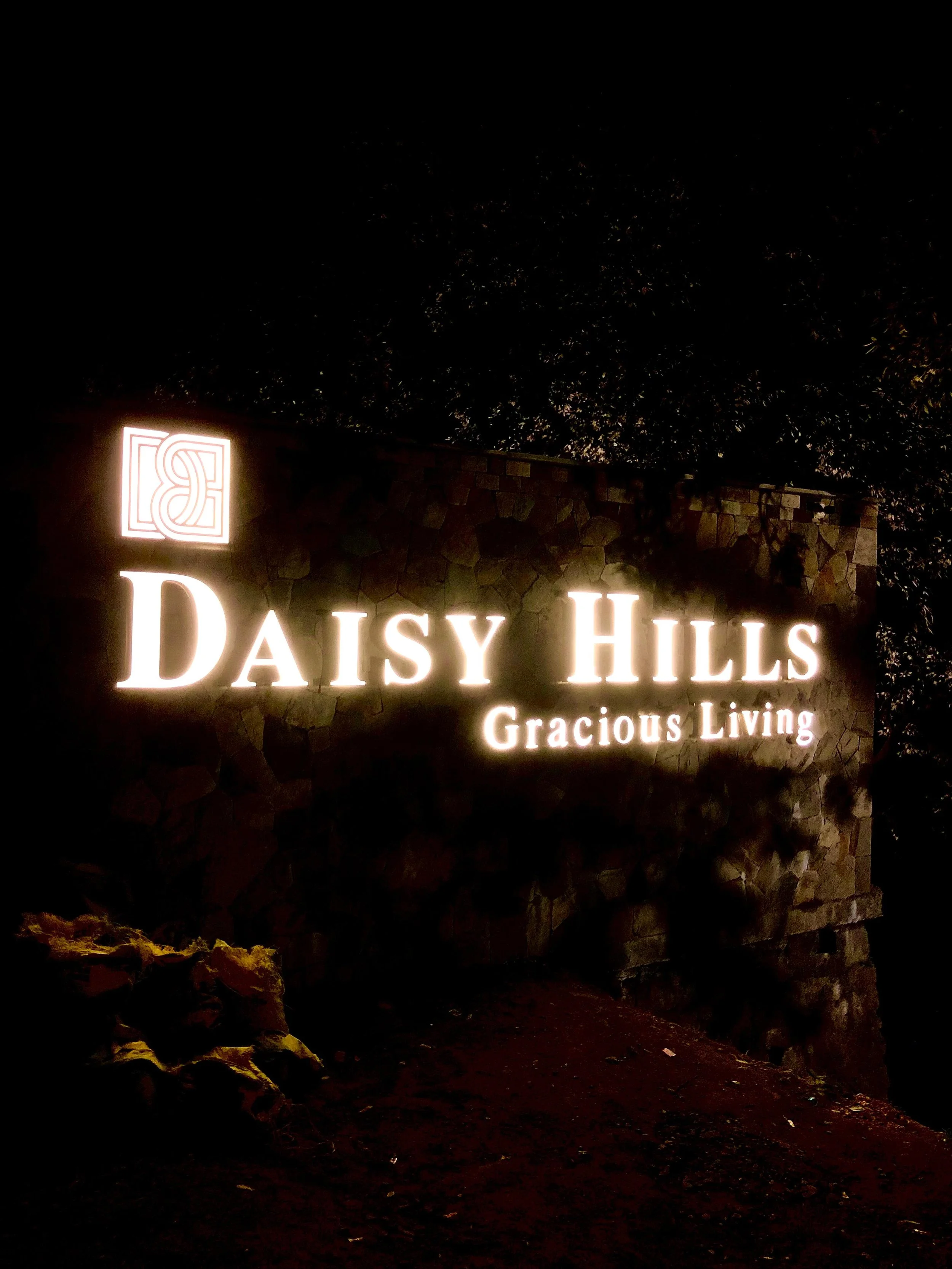 Illuminated sign reading "Daisy Hills Gracious Living" against a stone wall at night.