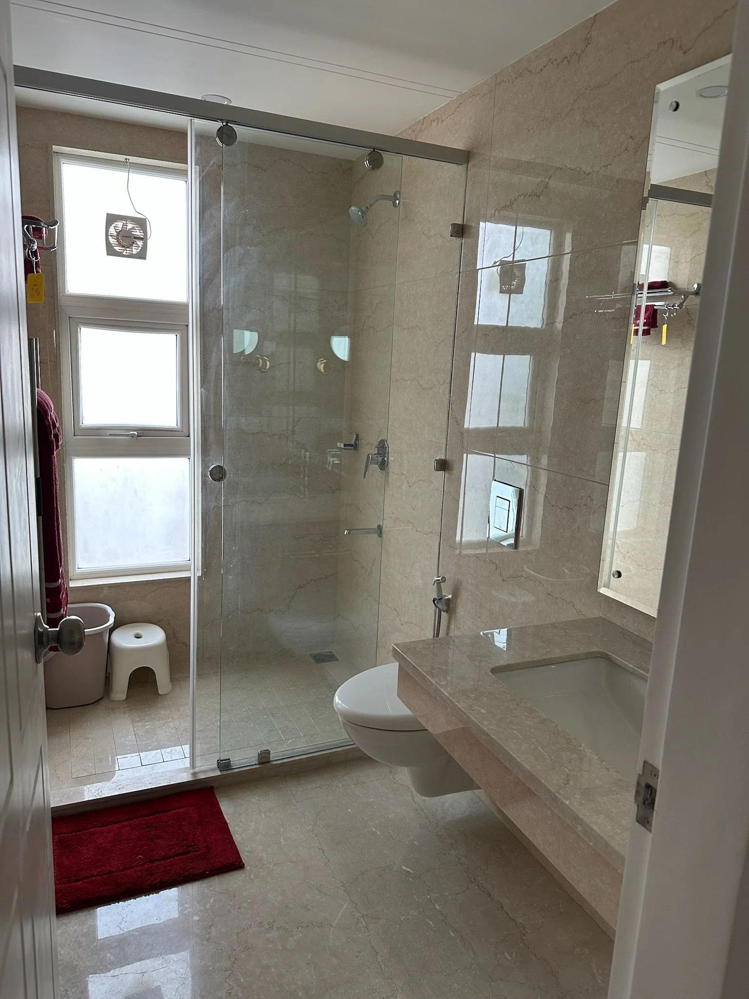 A bathroom with beige marble walls, a small sink with a marble countertop, and a large mirror. There are pink towels on a ledge and a door leading to a hallway with pink walls and a red rug.