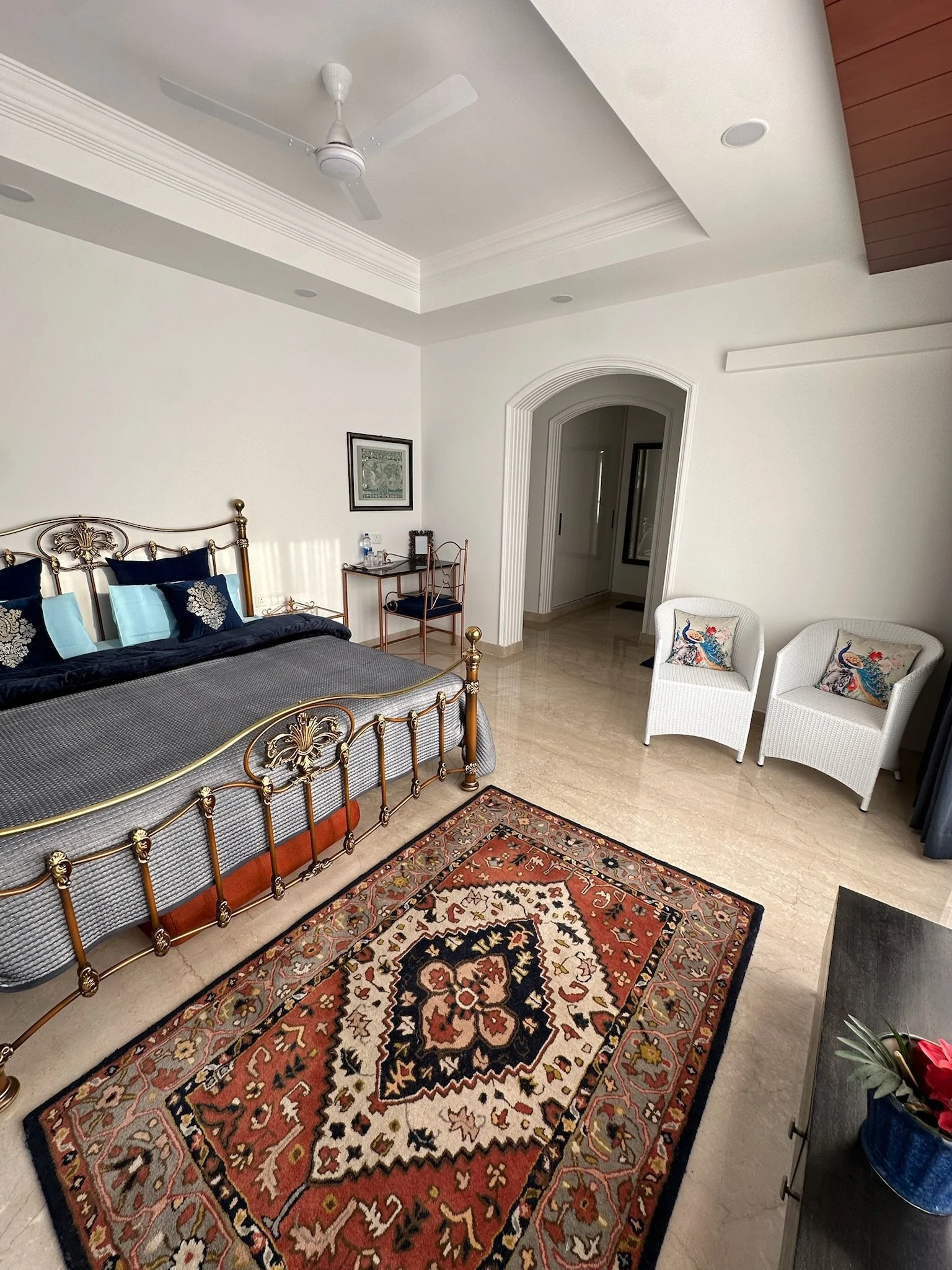 Living room with two beige armchairs with wooden frames, small side table, Persian-style rug, large window with cream curtains, outside snowy trees, marble floor, ceiling with recessed lighting.