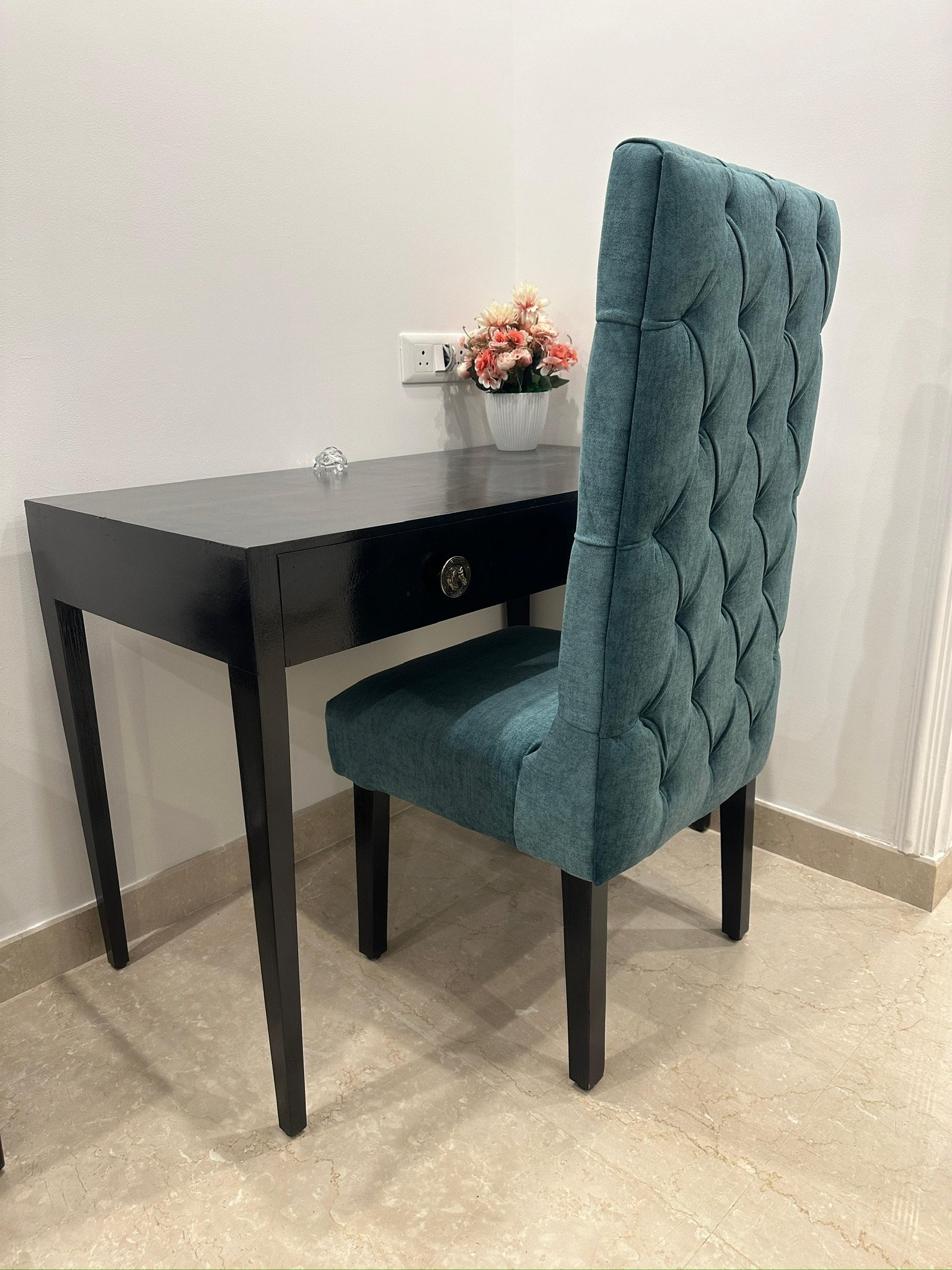 A black desk with a drawer, adorned with a potted pink and white artificial flower arrangement, next to a tall, teal upholstered high-back chair with button tufting, placed on a beige tiled floor against a white wall with an electrical outlet.