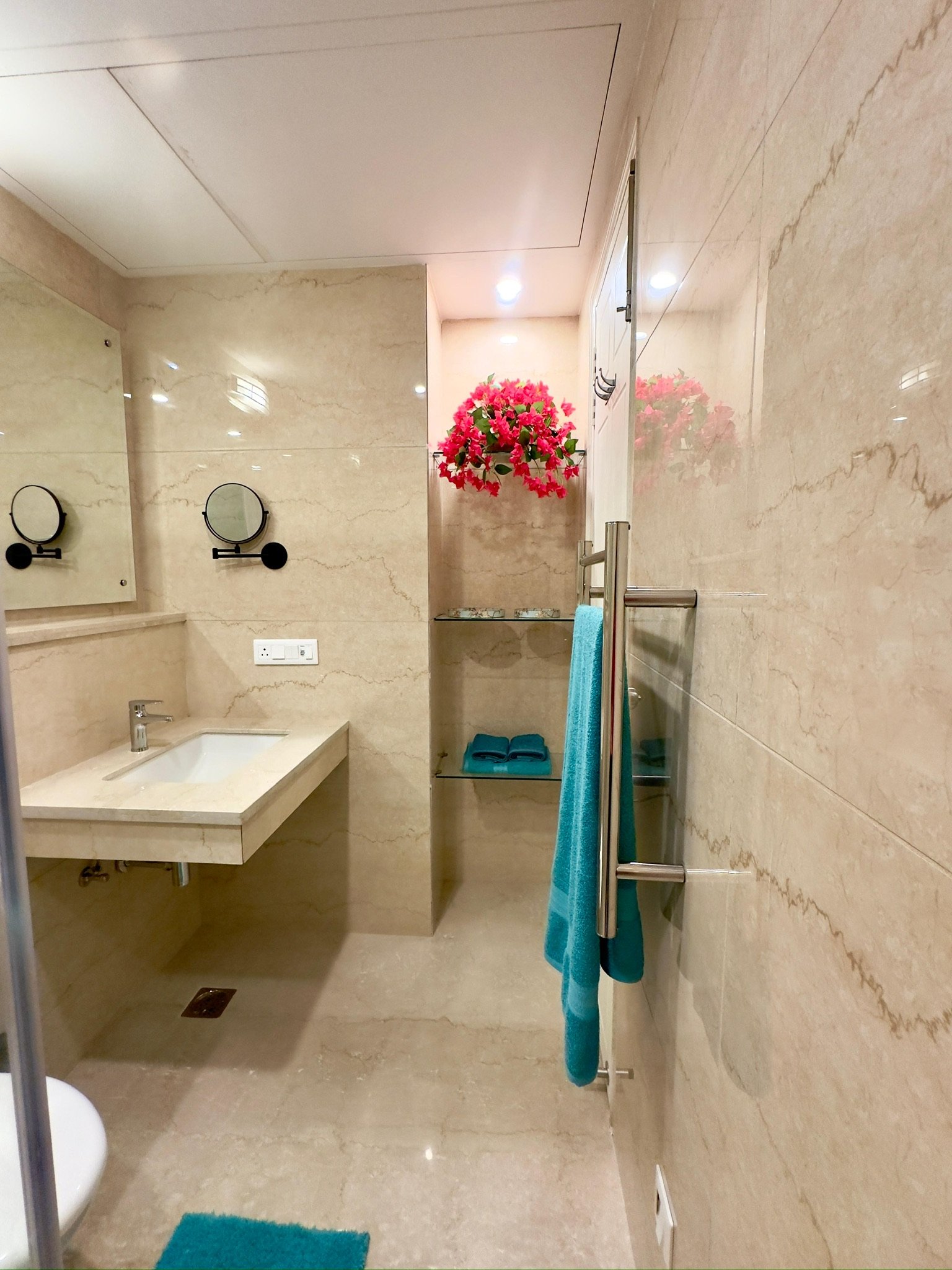A small bathroom with beige marble walls and floor, featuring a sink with a beige countertop, a large mirror, a small shelf with a black mirror, a towel rack with turquoise towels, a vibrant pink flower arrangement on the wall, and a teal rug on the floor.