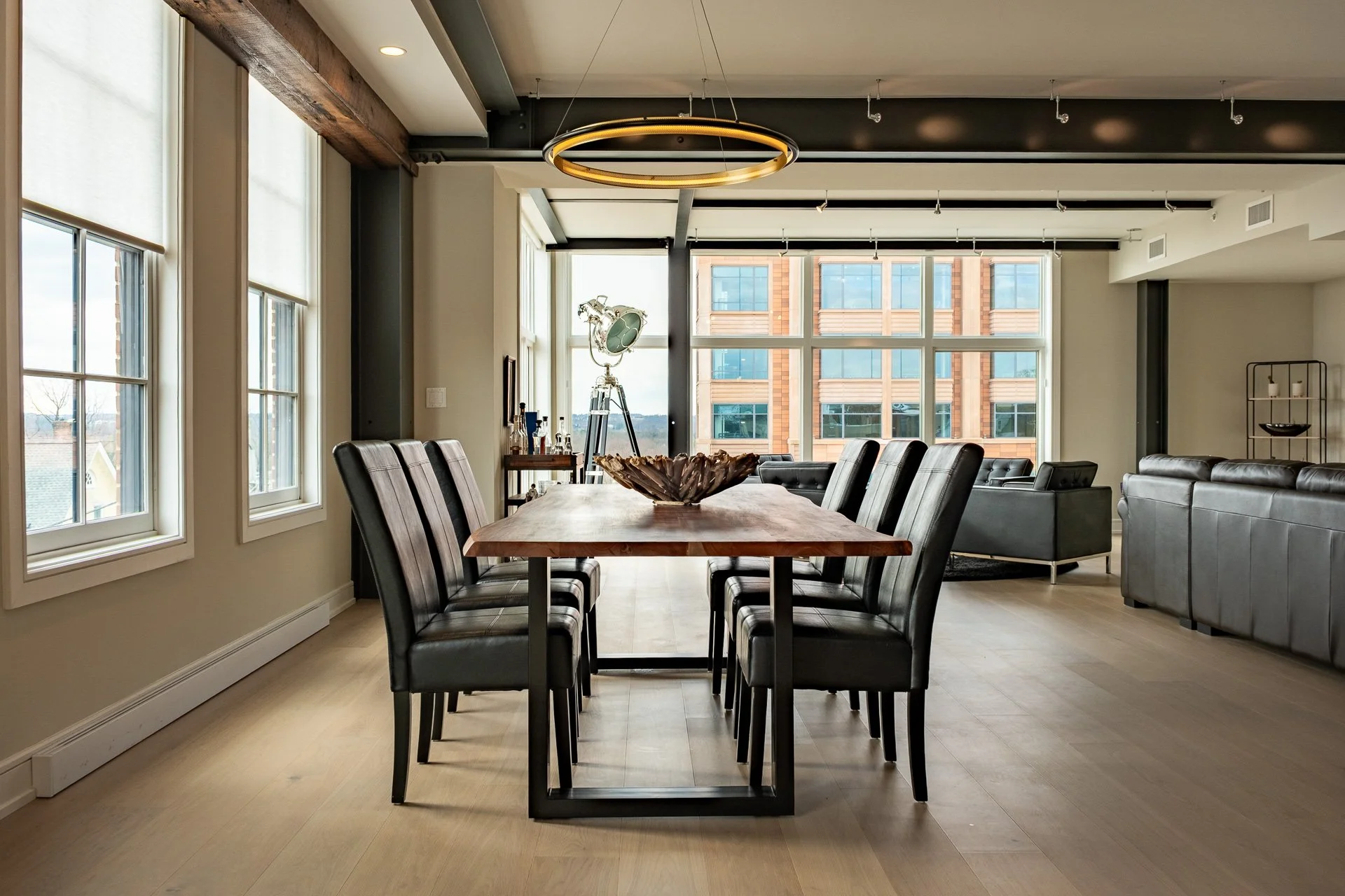 Modern dining and living area with a wooden table, black upholstered chairs, large windows, and additional seating in the background.