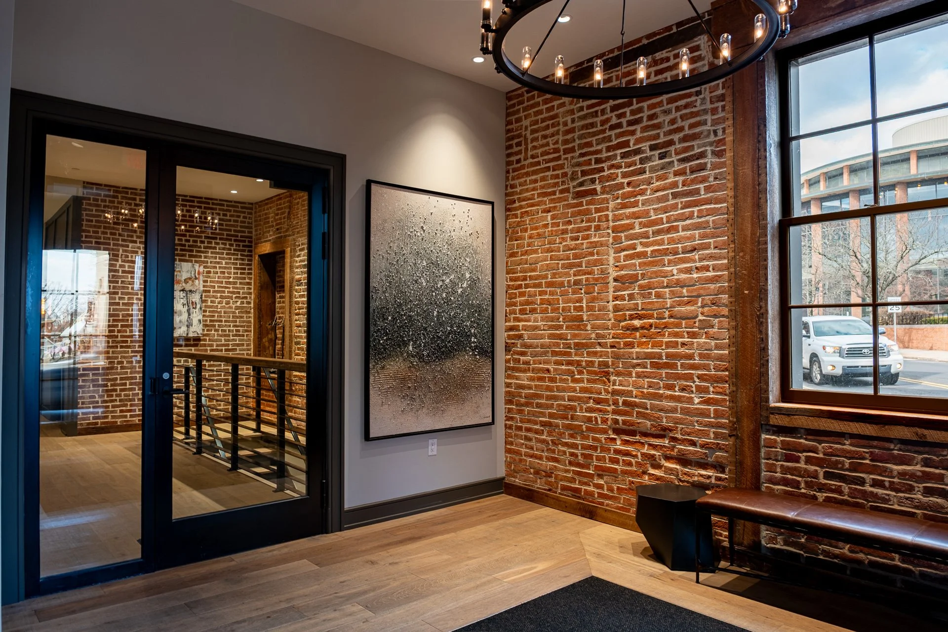 A modern interior with exposed red brick walls, large windows, and a glass door leading to a staircase. There is a large artwork on the wall, a black bench, and a circular chandelier hanging from the ceiling.