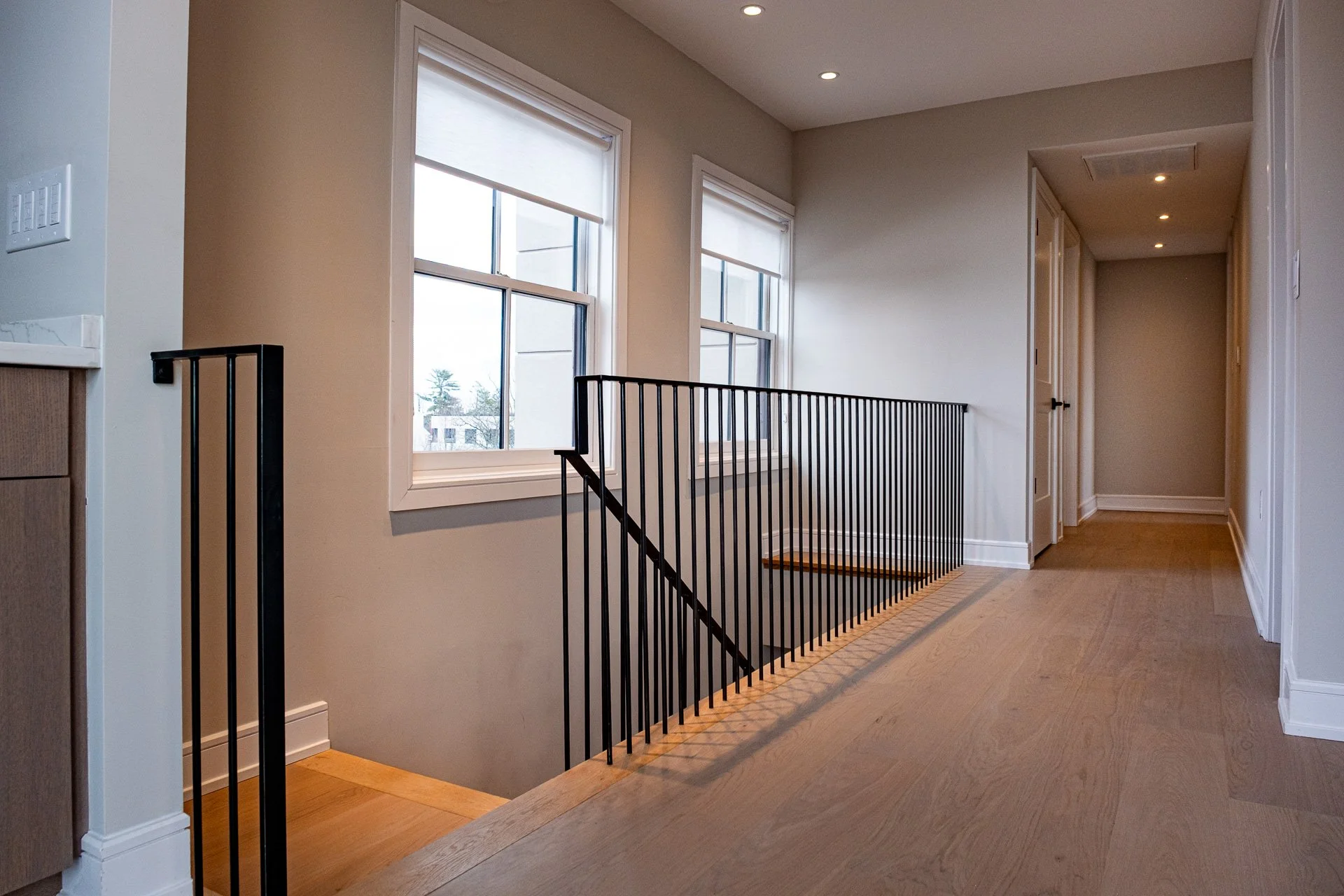 Bright hallway with wooden flooring, two large windows, and a staircase with black metal railing leading downstairs.