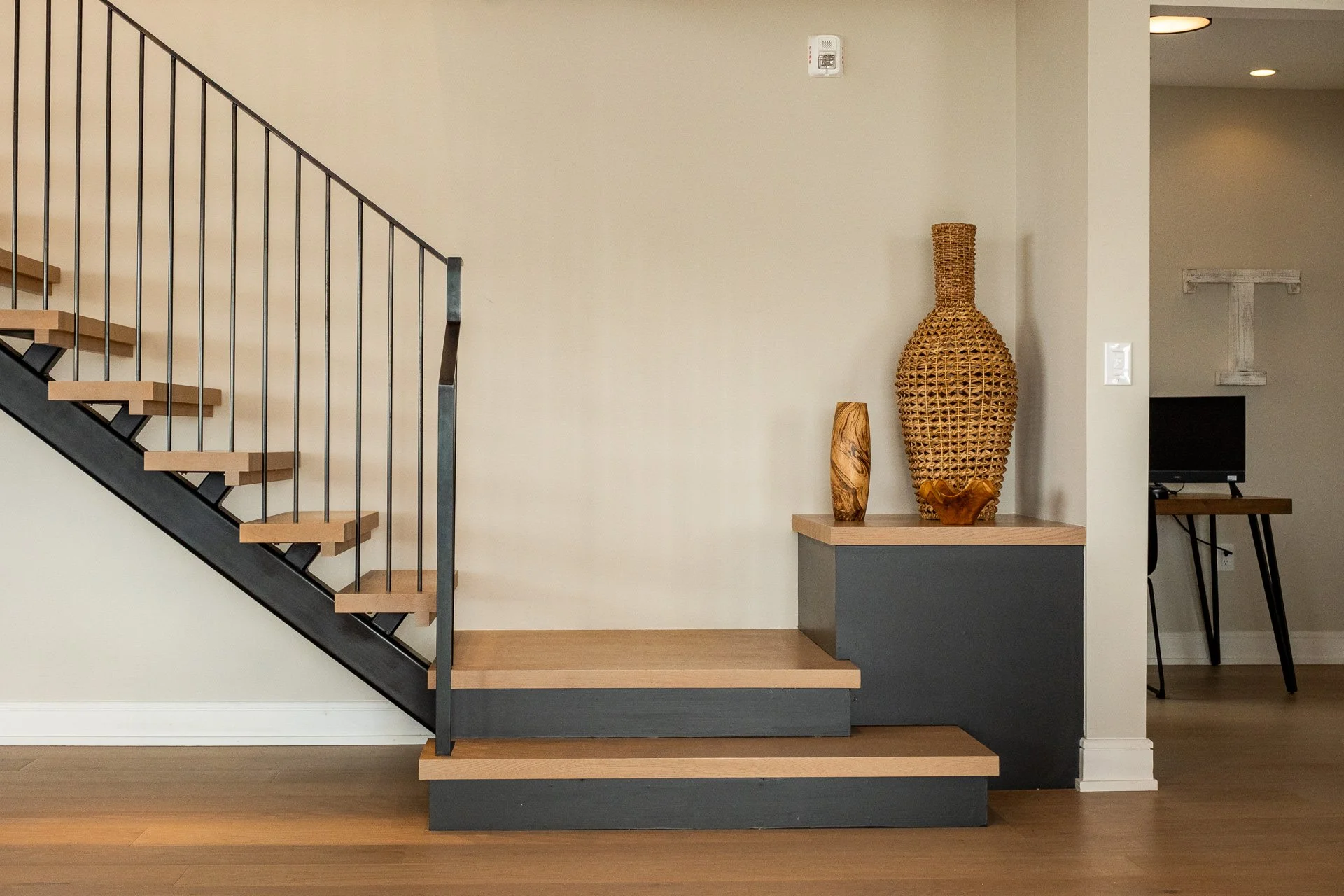 Interior of a home showing a staircase with wooden steps and black metal railings, decorated with three wooden vases and a large woven vase on a small platform, with a wall-mounted smoke detector and a doorway leading to a workspace with a computer a