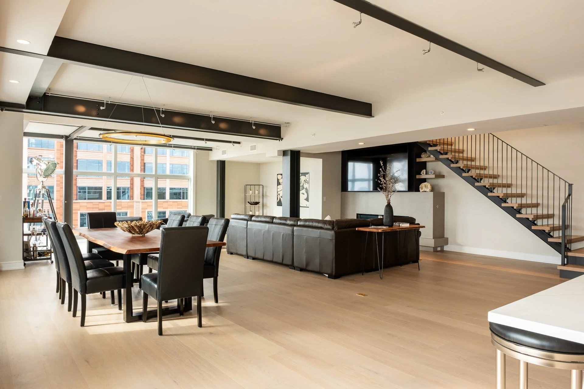 Modern dining and living room area with large windows, black leather dining chairs around a wooden table, a black leather sofa, staircase with wooden steps and black metal railing, decorative shelving, and a large flat-screen TV.