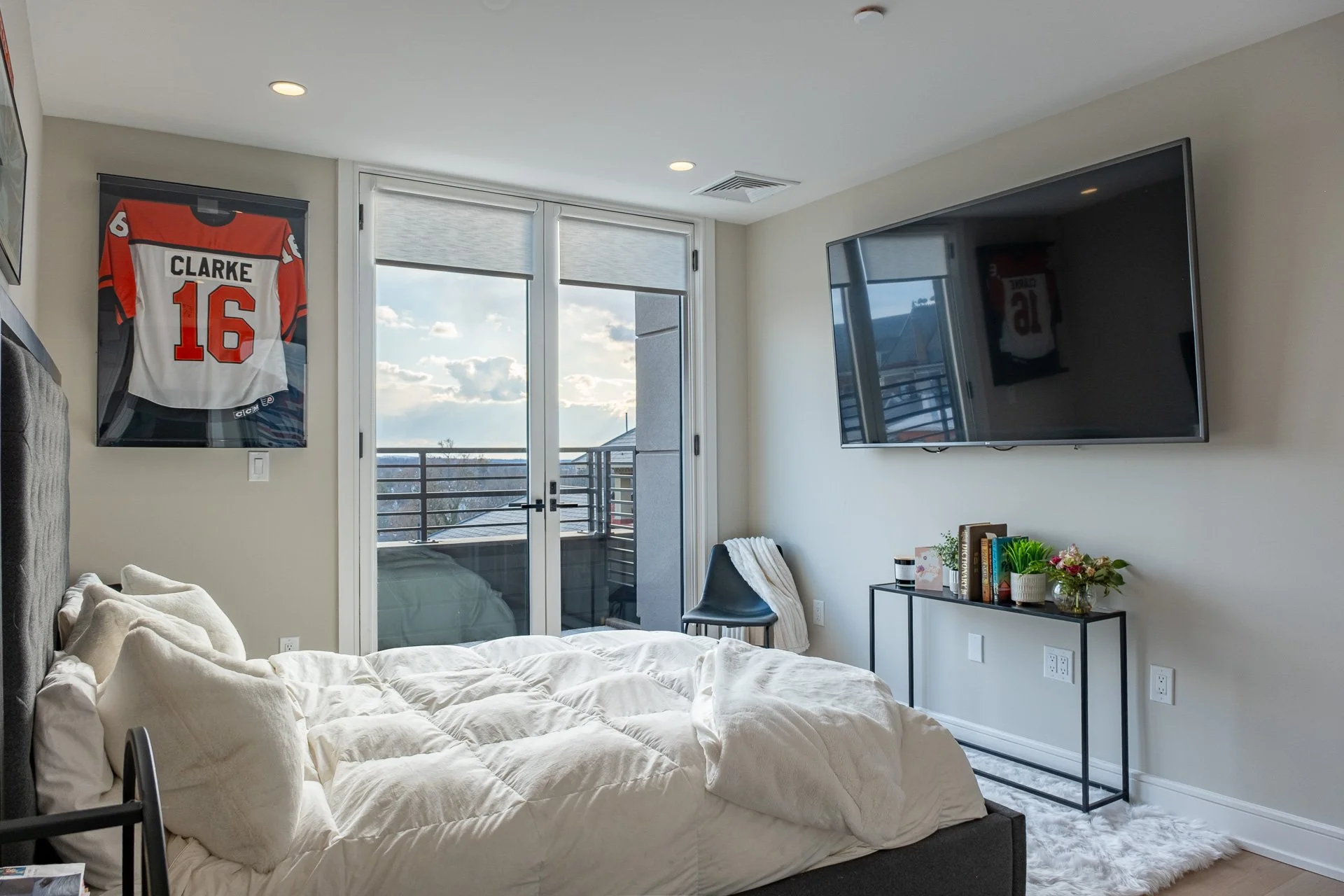 A modern bedroom with a neatly made bed, a mounted flat-screen TV, a black chair with a blanket, a small black table with books, plants, and decorations, glass door leading to a balcony showing an outdoor view with clouds, a framed sports jersey on t