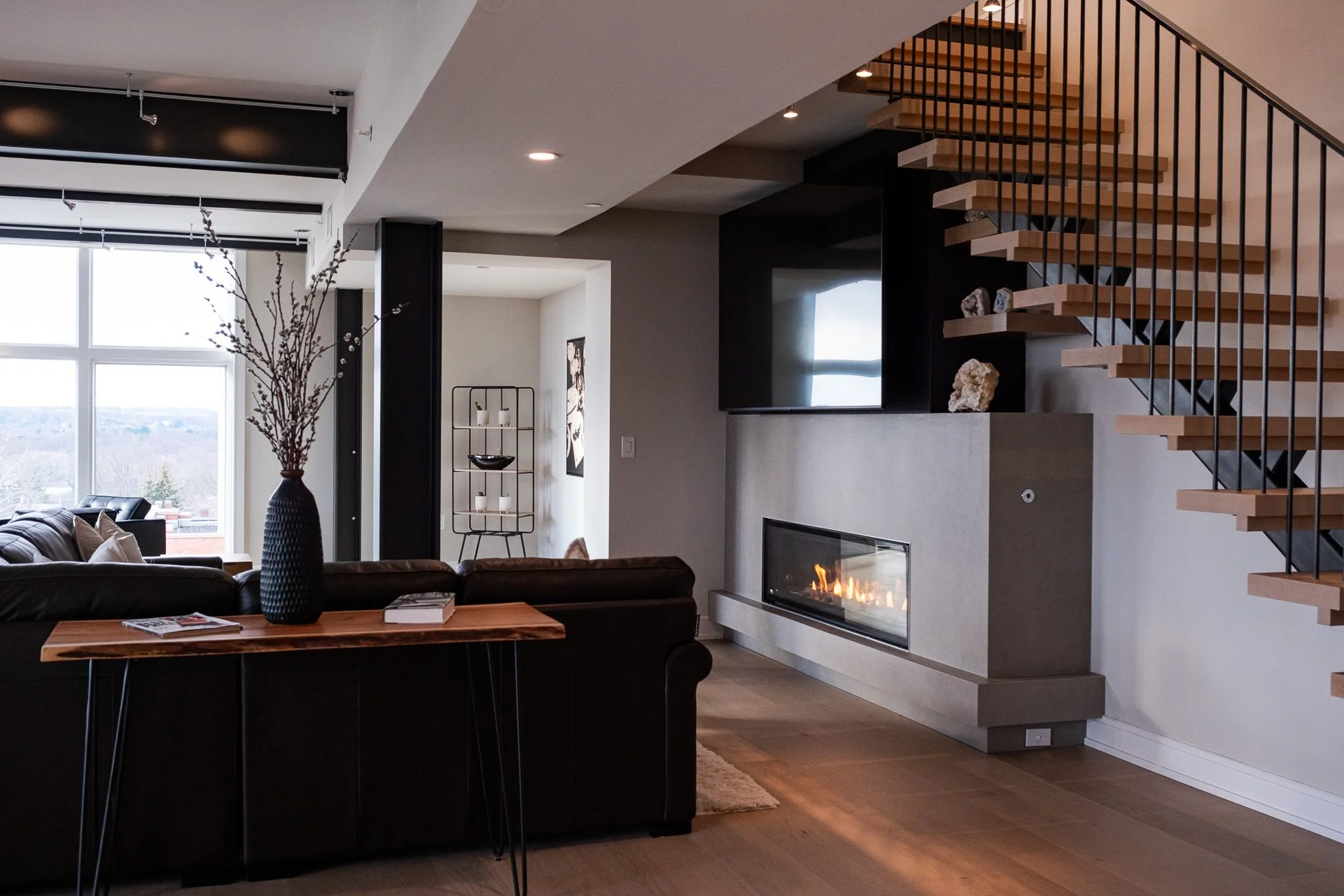 Modern living room with a large window, black sofa, wooden coffee table with a tall vase and books, fireplace, wall-mounted TV, staircase with wooden steps and black metal railing, and decorative items like a art piece and a shelving unit.