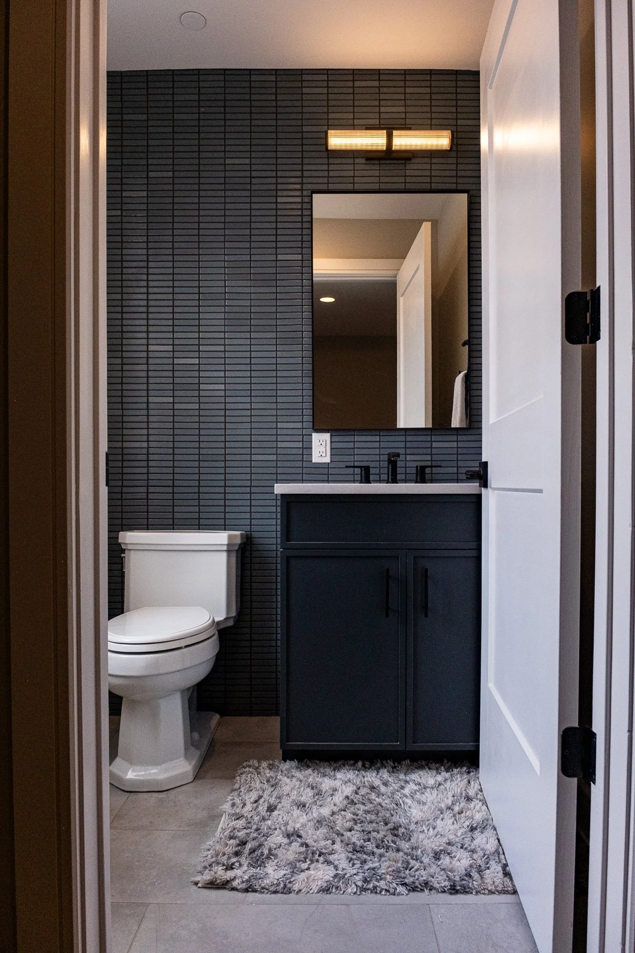 A modern bathroom with a white toilet, a dark blue vanity with a white countertop, and a large mirror. The wall behind the vanity is covered with small horizontal black tiles. There is a rectangular light fixture above the mirror and a fluffy gray rug on the floor.