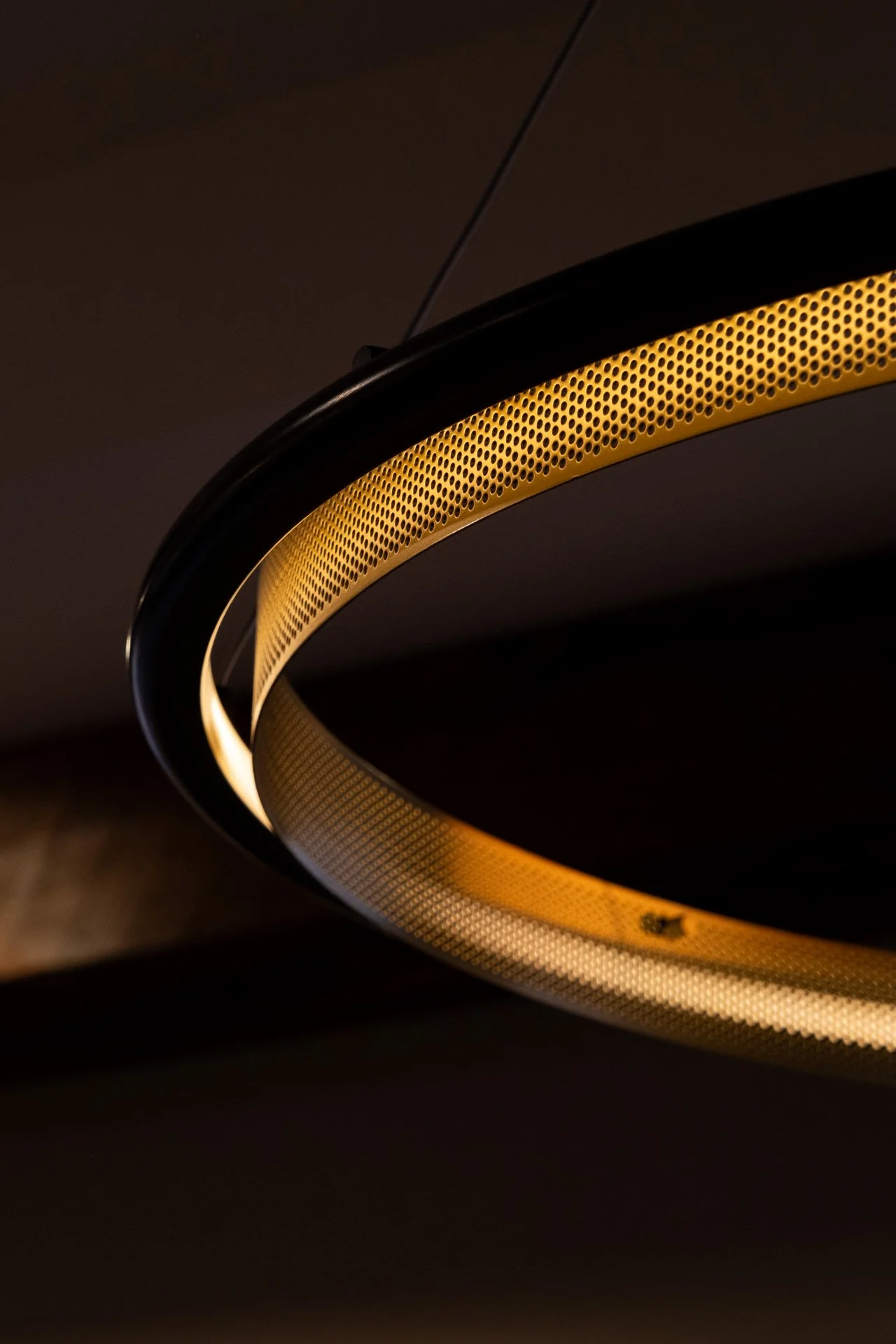 Close-up of a modern, circular ceiling light fixture with a black outer ring and a perforated gold inner ring, illuminated in a dark room.