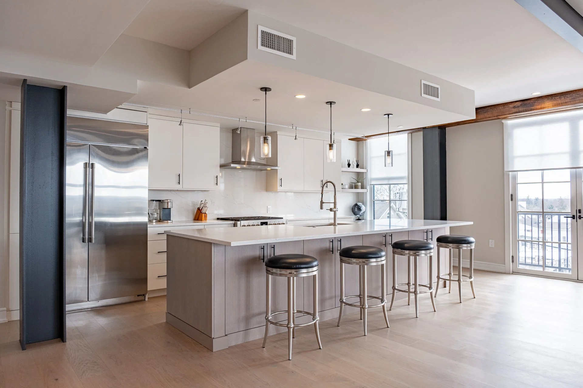 Modern kitchen with white cabinets, stainless steel appliances, and an island with four black and metallic bar stools, natural light from large windows and a sliding glass door.
