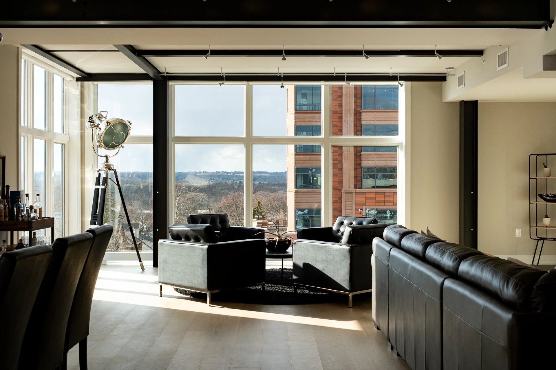 Modern living room with large windows, black leather sofas, a vintage floor lamp, and a view of city buildings and sky outside