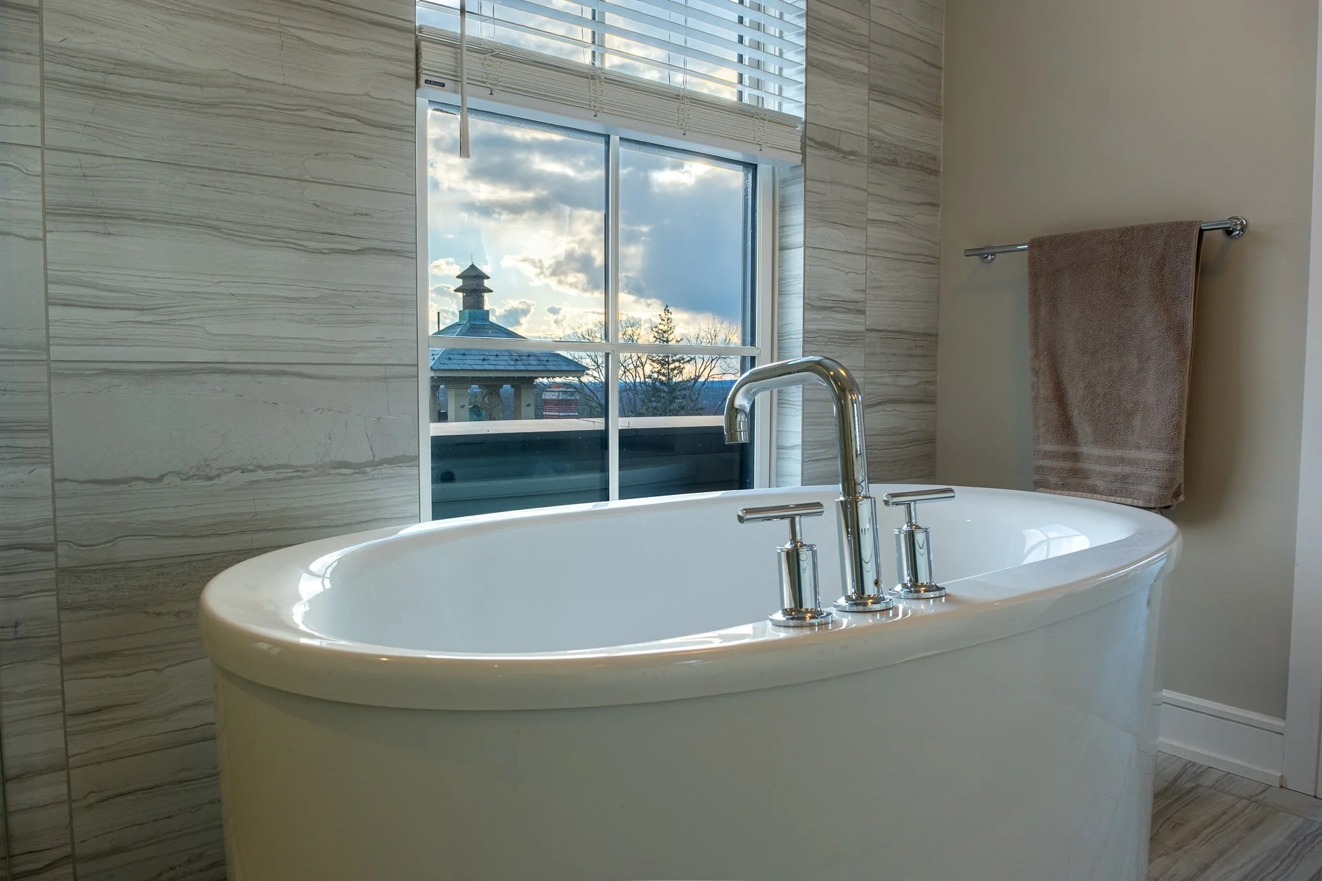 A modern bathroom with a white bathtub and a large window showing an outdoor scene with a roof and a tower. There is a brown towel hanging on a silver towel rack on the wall.