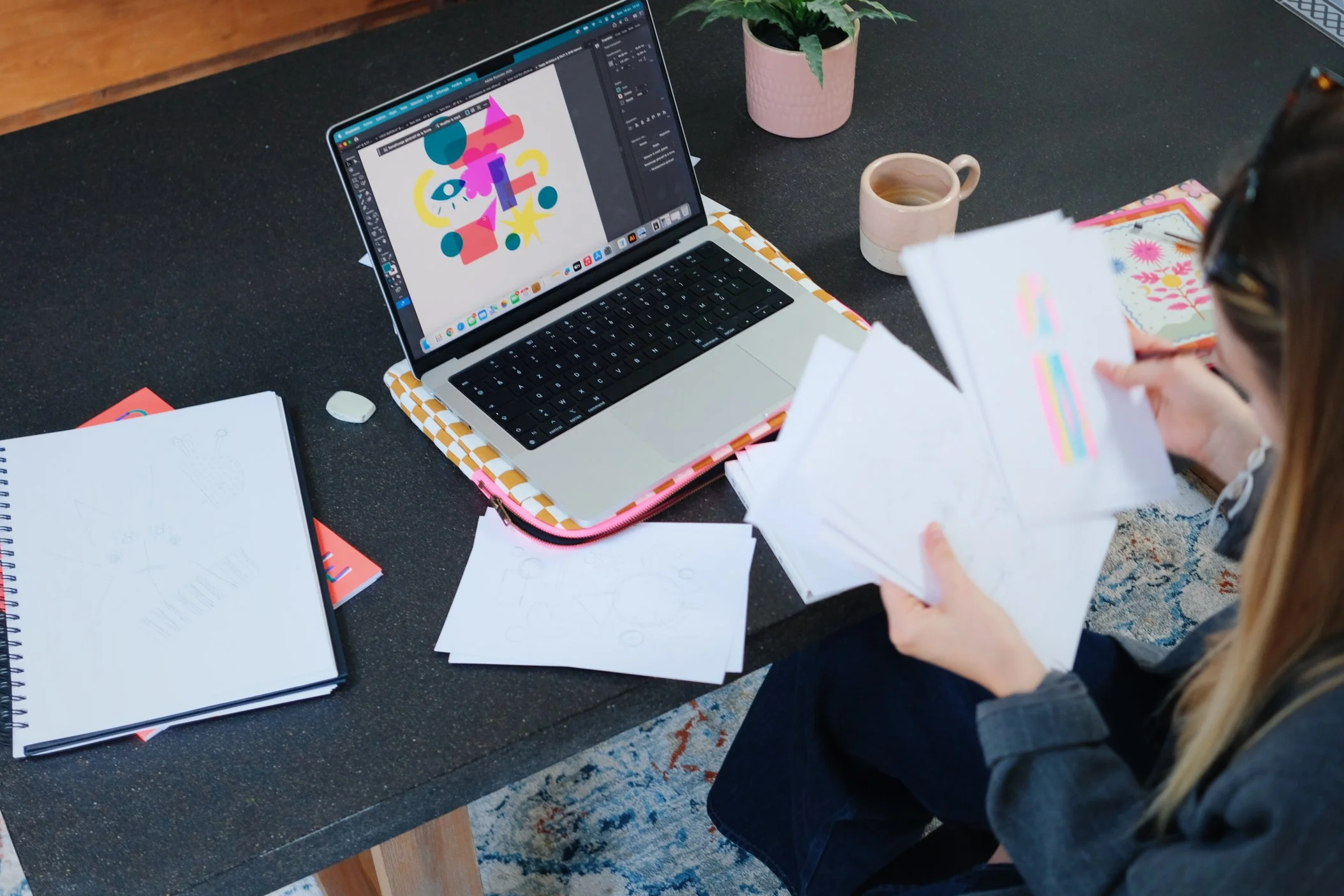 Personne assise à un bureau, utilisant un ordinateur portable avec un fond coloré de dessin numérique sur l'écran, entourée de papiers, livres et une tasse à café, avec une plante en pot à côté.
