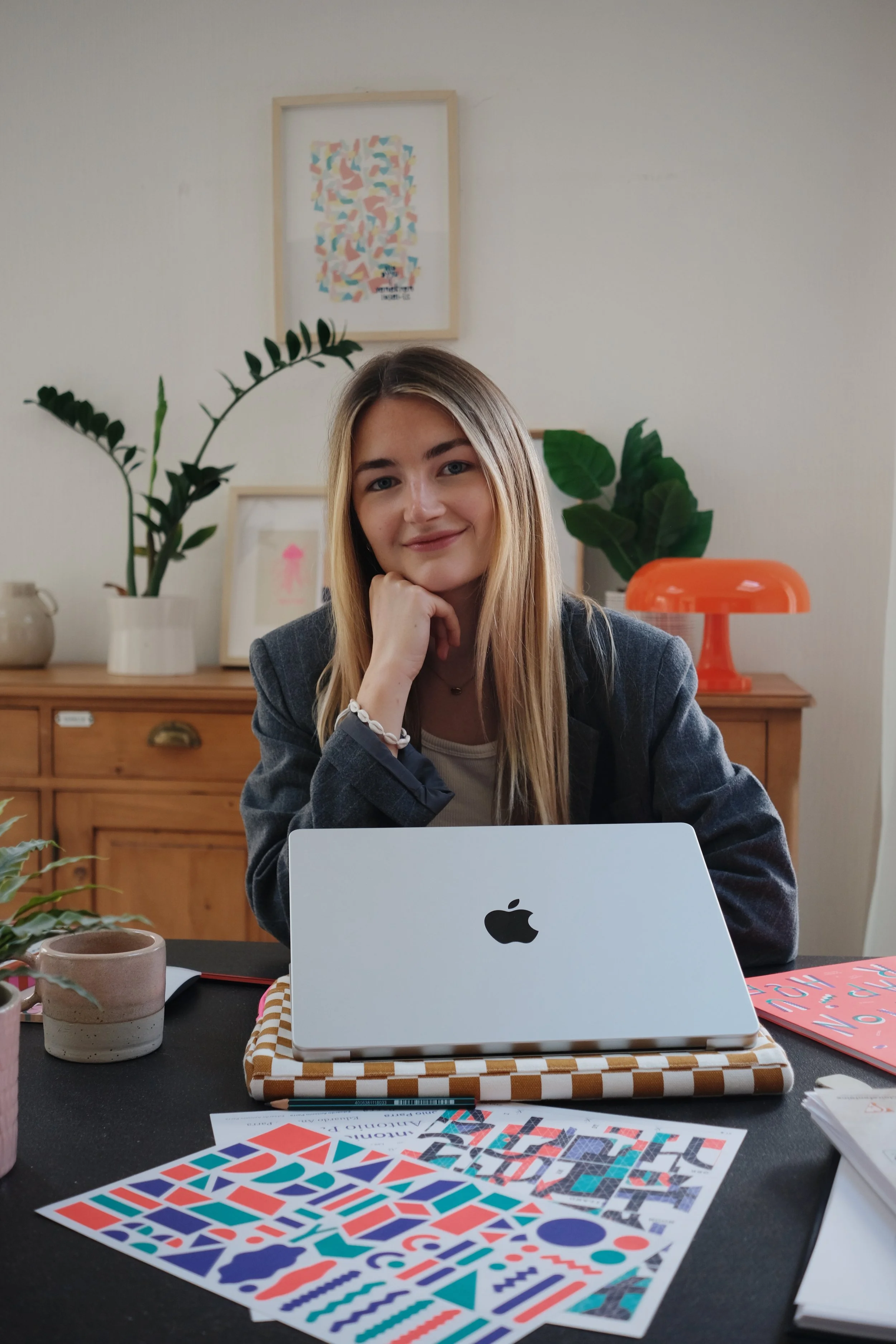 Une jeune femme avec des cheveux longs blonds, portant un blazer gris, assise à une table avec un ordinateur portable Apple. La table a des affiches colorées et des magazines. En arrière-plan, il y a des plantes vertes, un tableau avec une œuvre abstraite et un objet orange en forme de lampe ou de décor. La pièce est lumineuse avec un mur blanc.