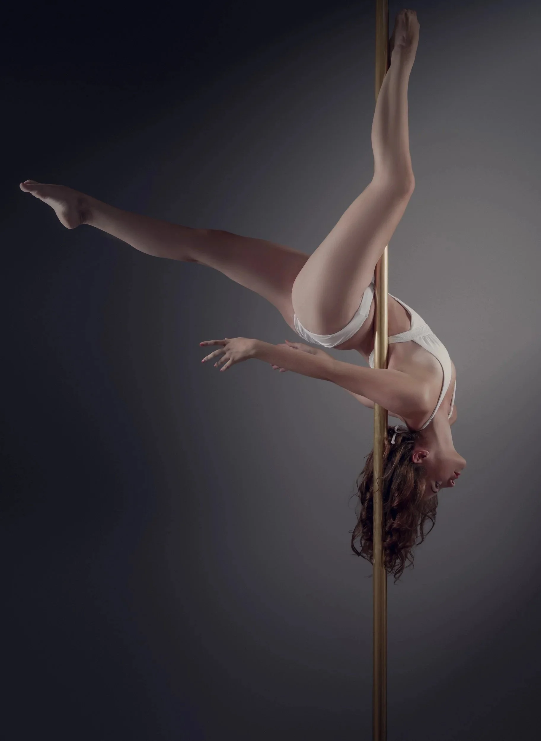 A woman performing a pole dance upside down, balancing with her legs gripping the pole and arms extended, in a dimly lit studio.