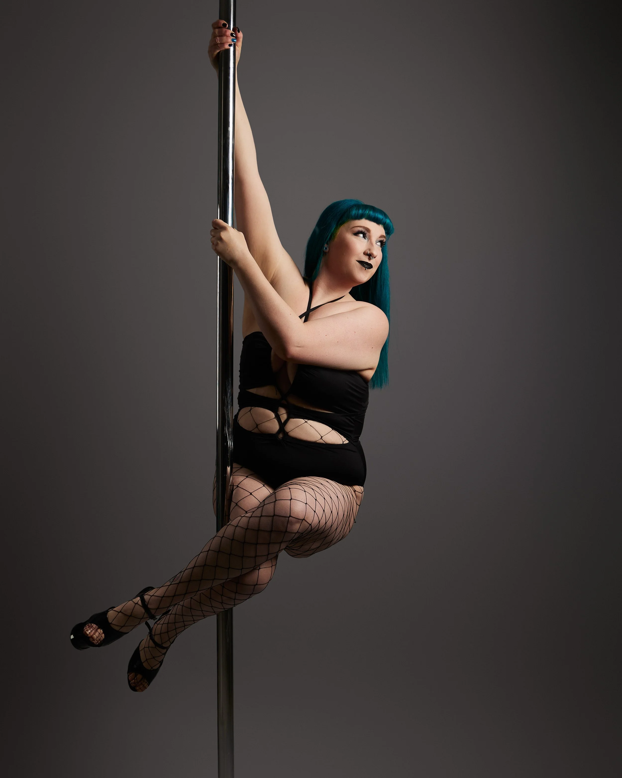 A woman with blue hair and dark attire performing a pole dance, sitting on a vertical pole against a gray background.
