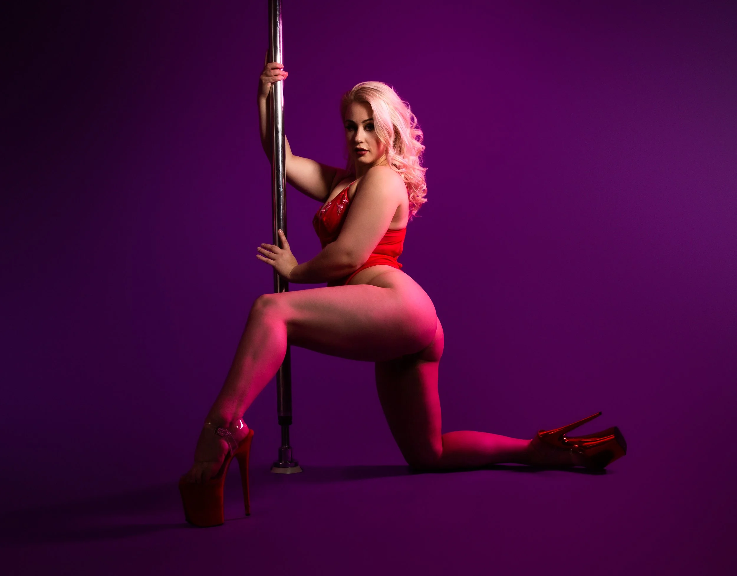 A woman with pink hair wearing red lingerie and high heels, posed on a purple background, holding a pole.