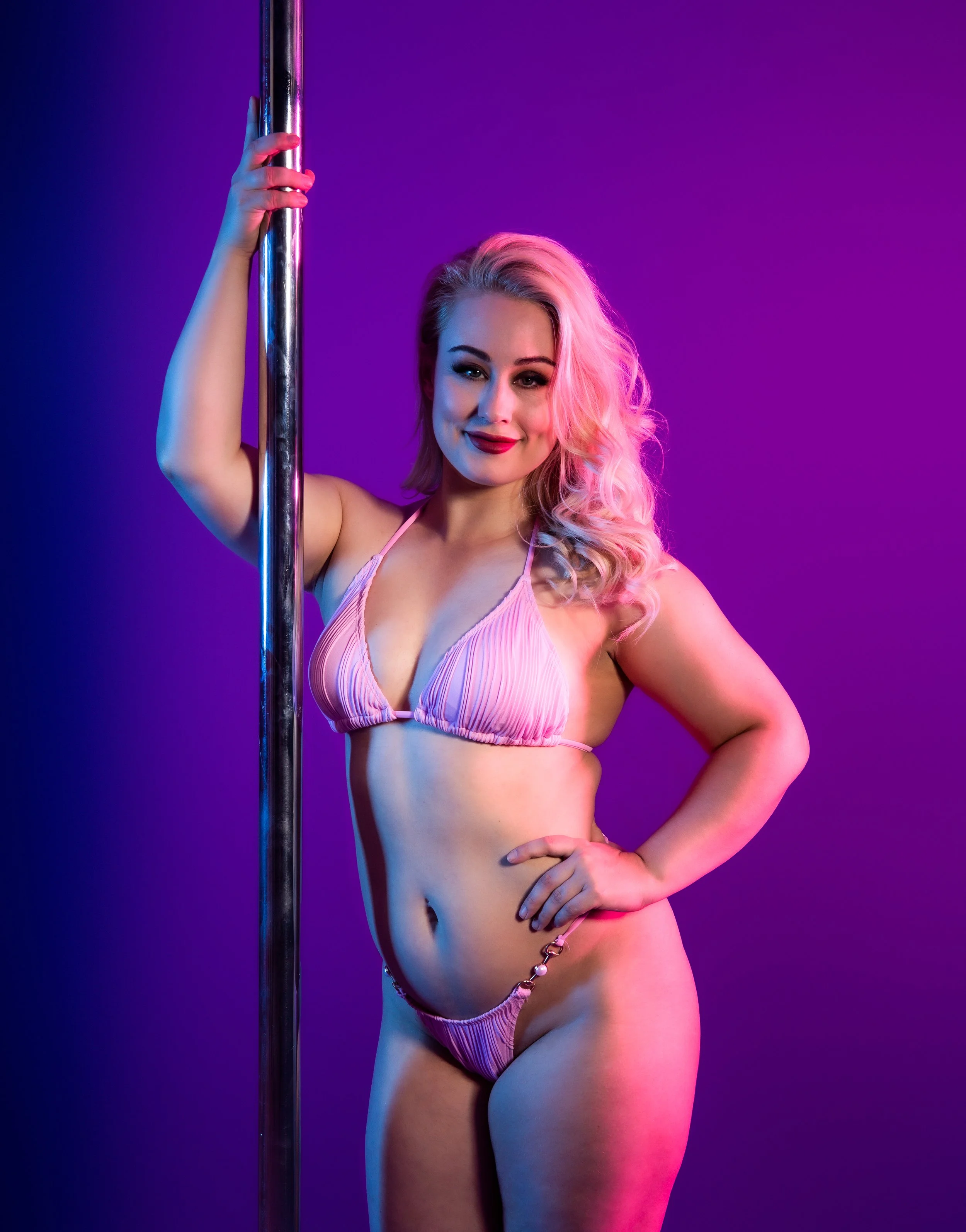 A woman with blonde hair in a pink striped bikini posing with a pole against a purple background.