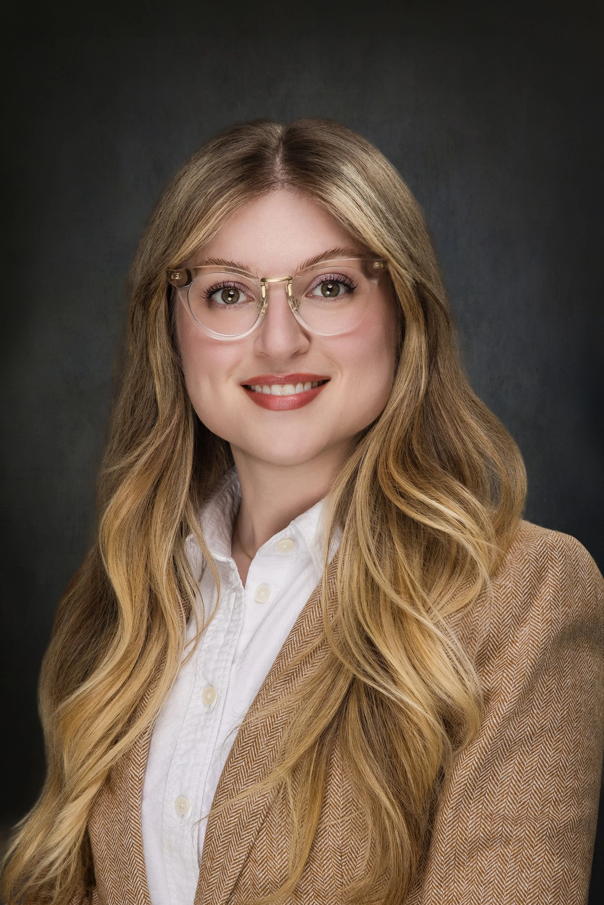 A woman with long wavy blonde hair, wearing clear-framed glasses, a white button-up shirt, and a brown blazer, smiling against a dark background.