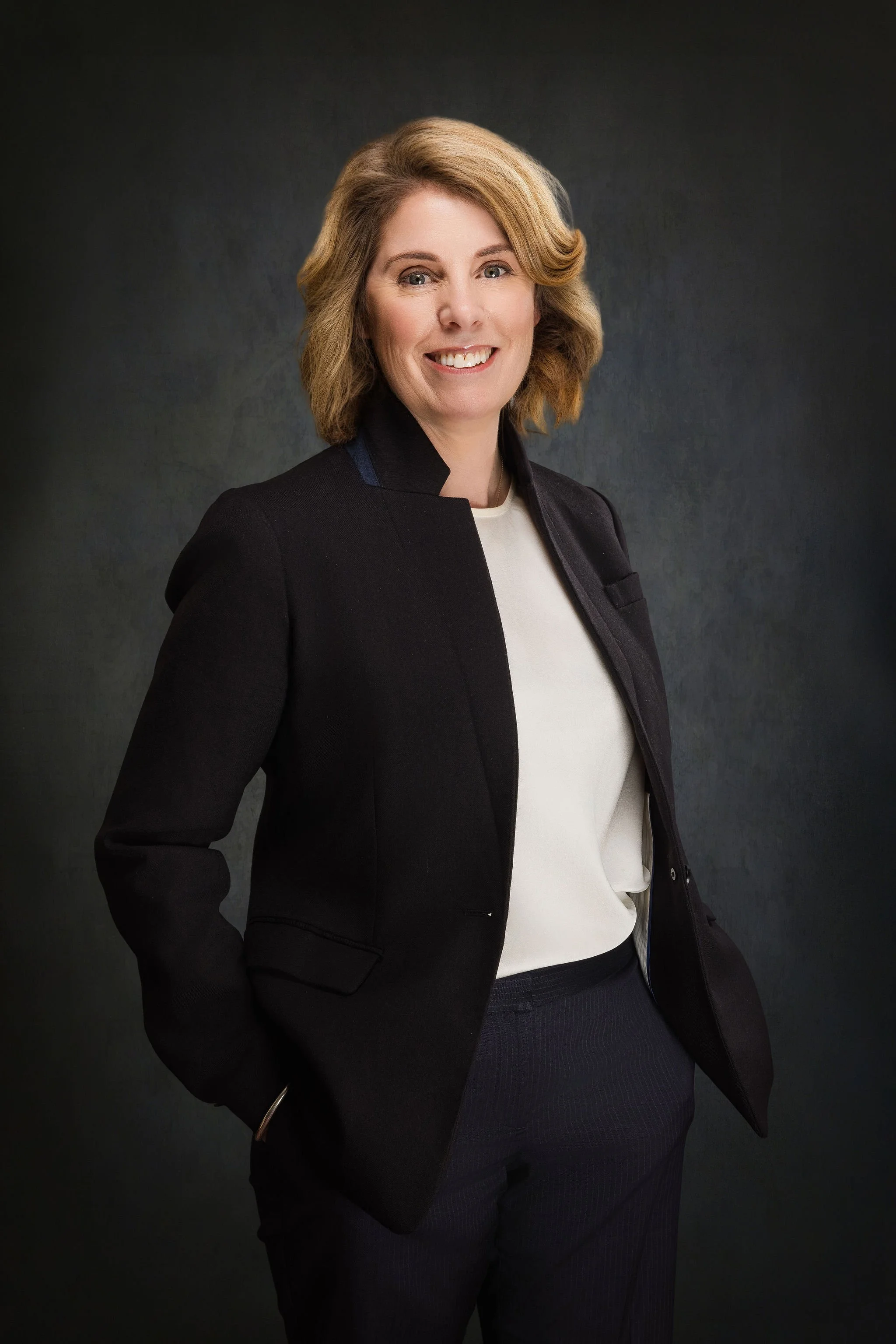 A woman with shoulder-length blonde hair wearing a black blazer over a white top, posing in front of a dark background.