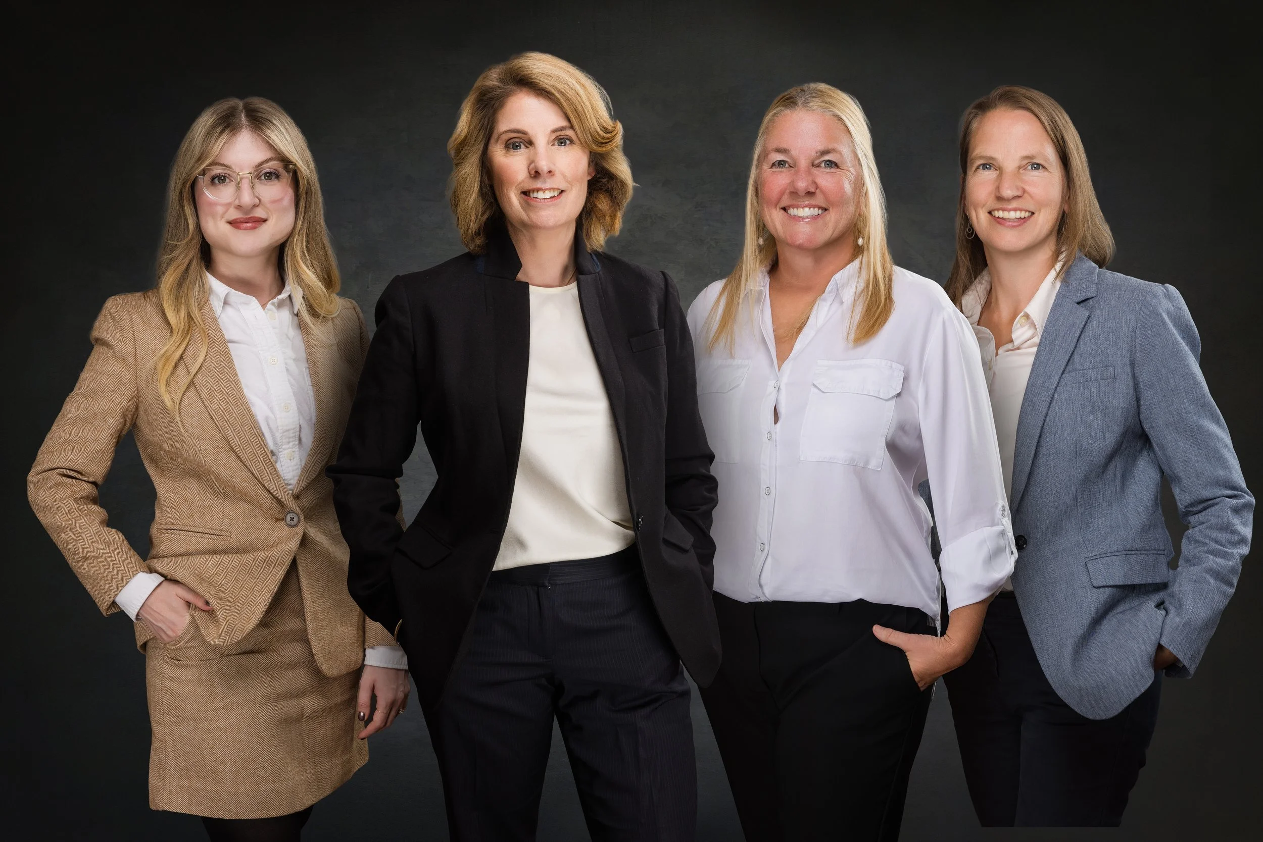 Four women standing together in business attire against a dark background, smiling at the camera.