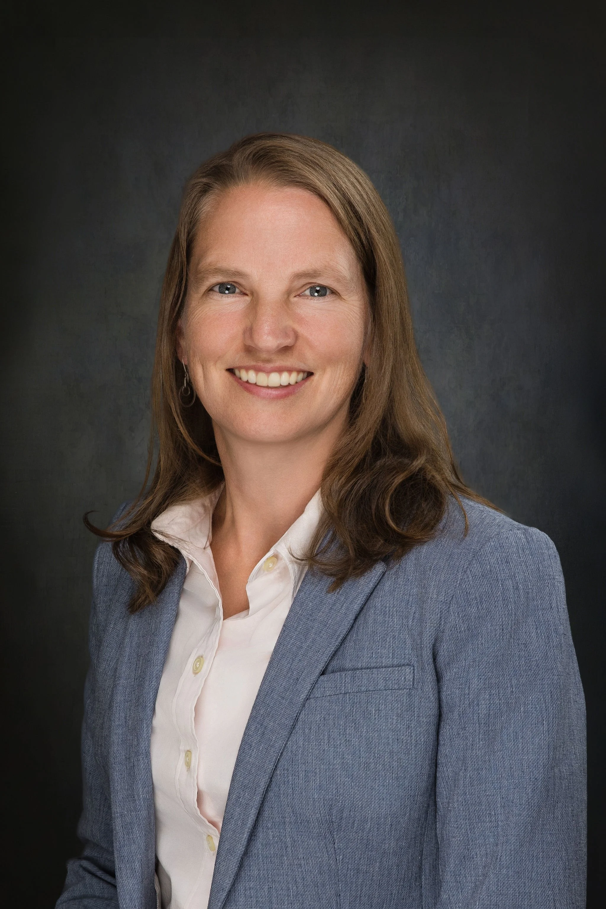 A professional woman with shoulder-length brown hair, wearing a light gray blazer and a white collared shirt, smiling against a dark background.