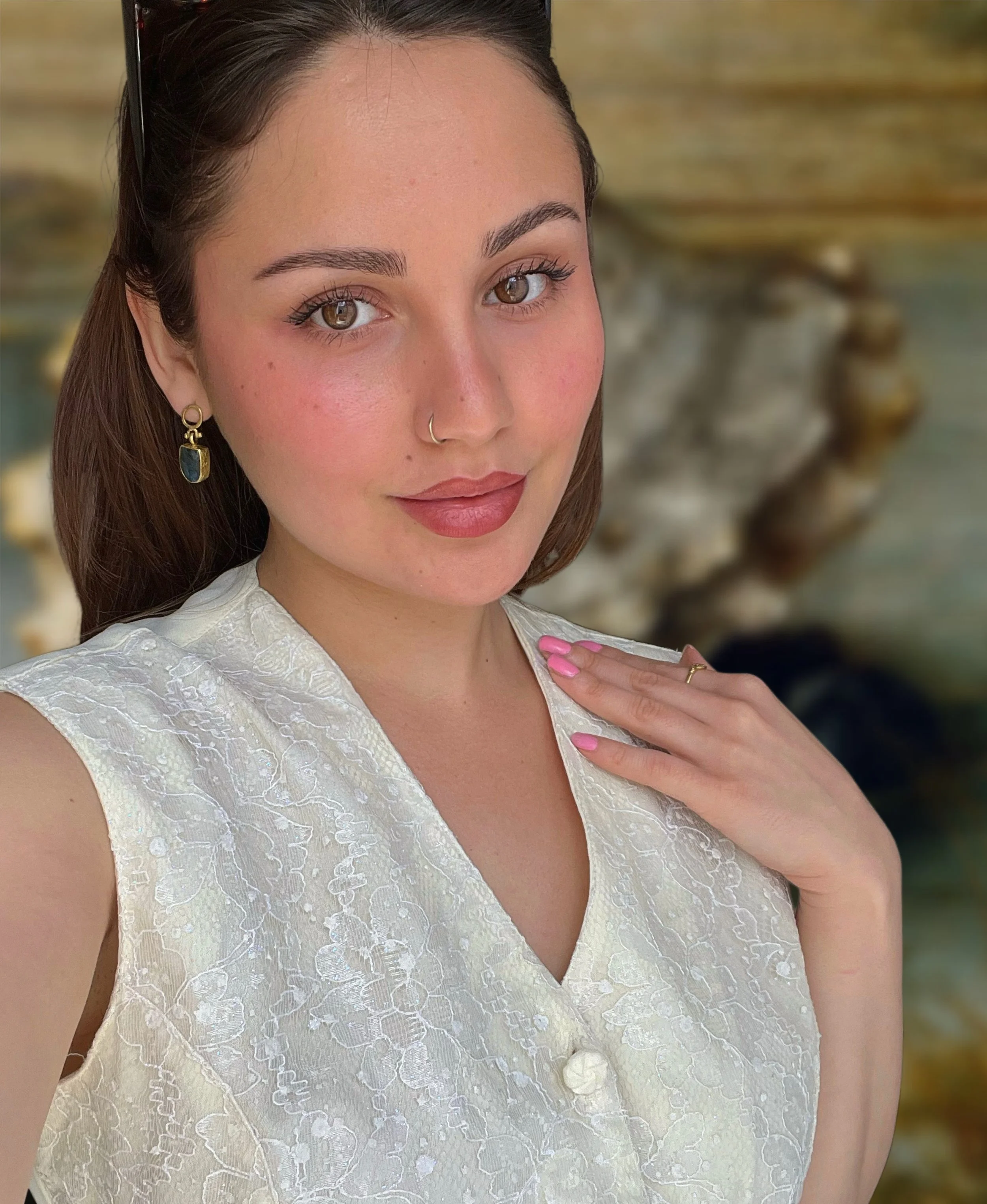A young woman with brown hair, brown eyes, and light makeup, wearing a white lace dress, a small hoop nose ring, and a gold earring with a dark stone. She is touching her collarbone with her right hand, which has pink nail polish, and is outdoors in front of a blurred background.