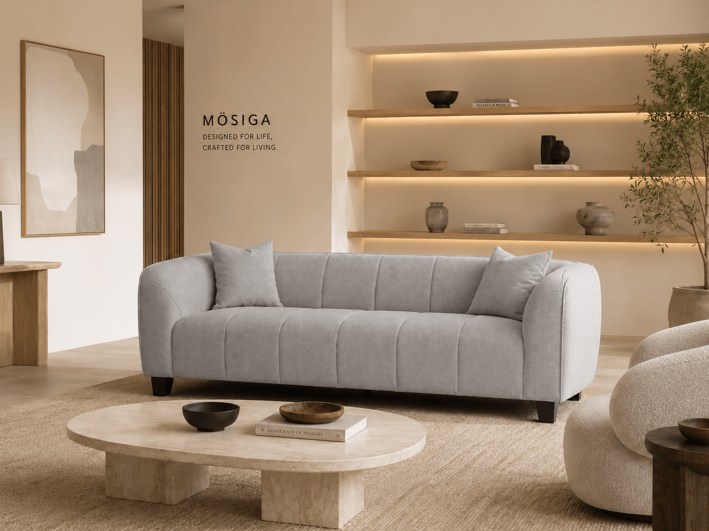 A modern, minimalist living room with a light gray sofa, a round marble coffee table holding small bowls and books, beige textured armchairs, a beige rug, and a built-in wooden shelf with decorative vases and books. There is a large potted plant on t