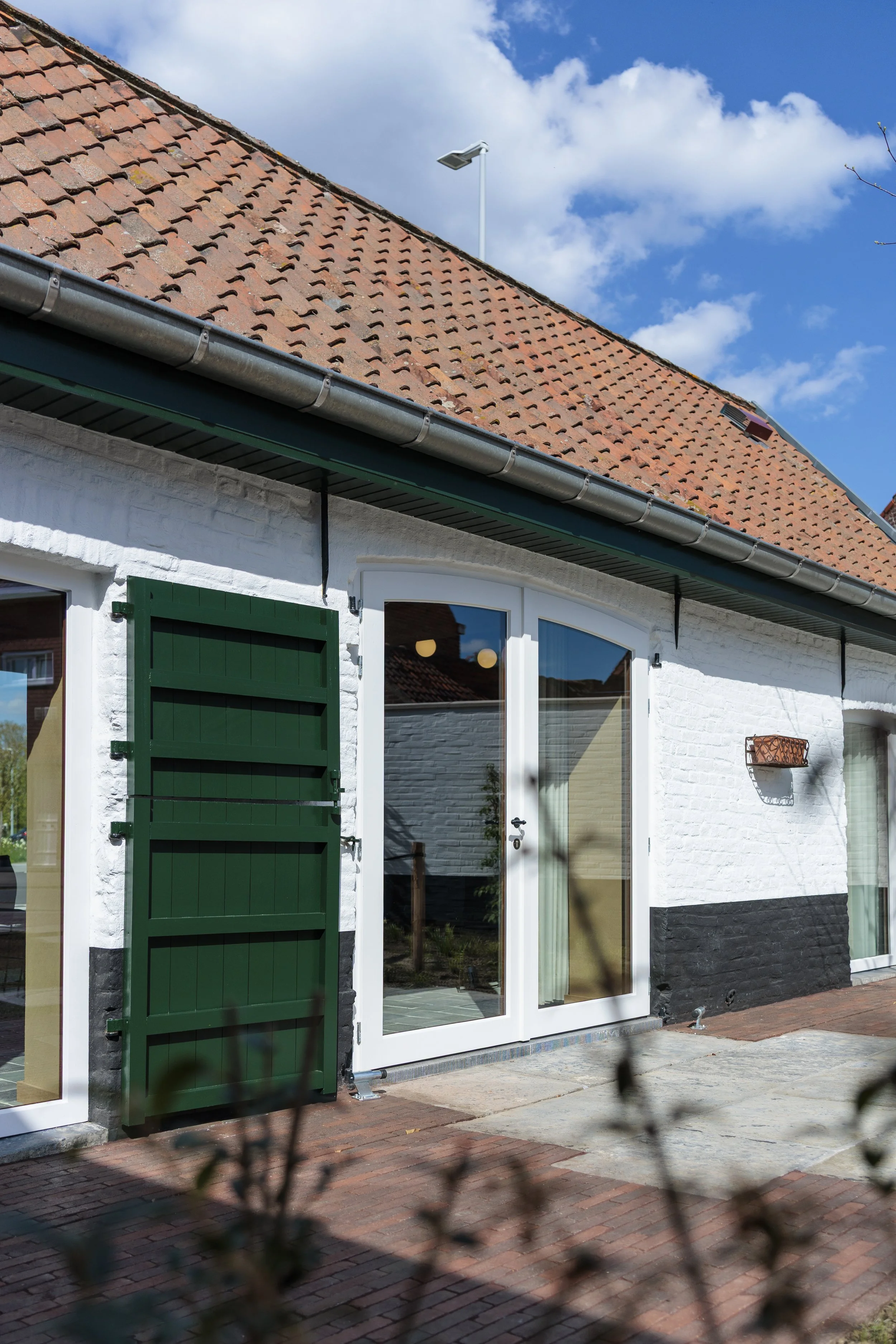 De afbeelding toont de gevel van een wit huis met grote ramen en een groene luikdeur. Het huis heeft een rood dak met dakpannen en er is een buitenlamp en een plantenbak aan de muur. De sky is blauw met witte wolken.