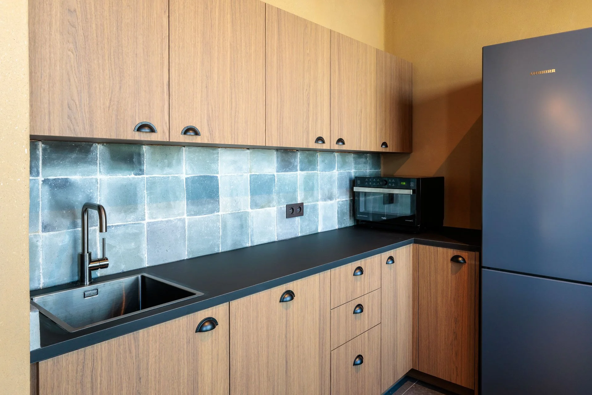 Een moderne keuken met houten kasten, een zwart werkblad, een gootsteen, een zwarte magnetron en een grote koelkasten. De muur achter het werkblad is bedekt met groene bakstenen.