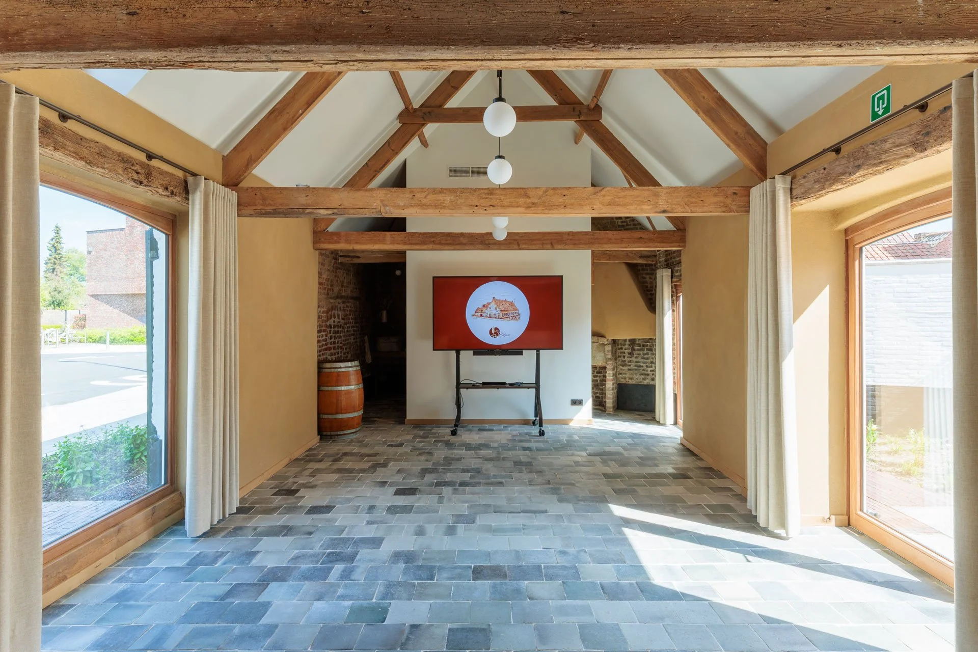 Interieur van een ruimte met grote ramen, houten balken en een tv-scherm met een logo van een gebouw en tekst onderaan, vloer met gevlamde tegels.