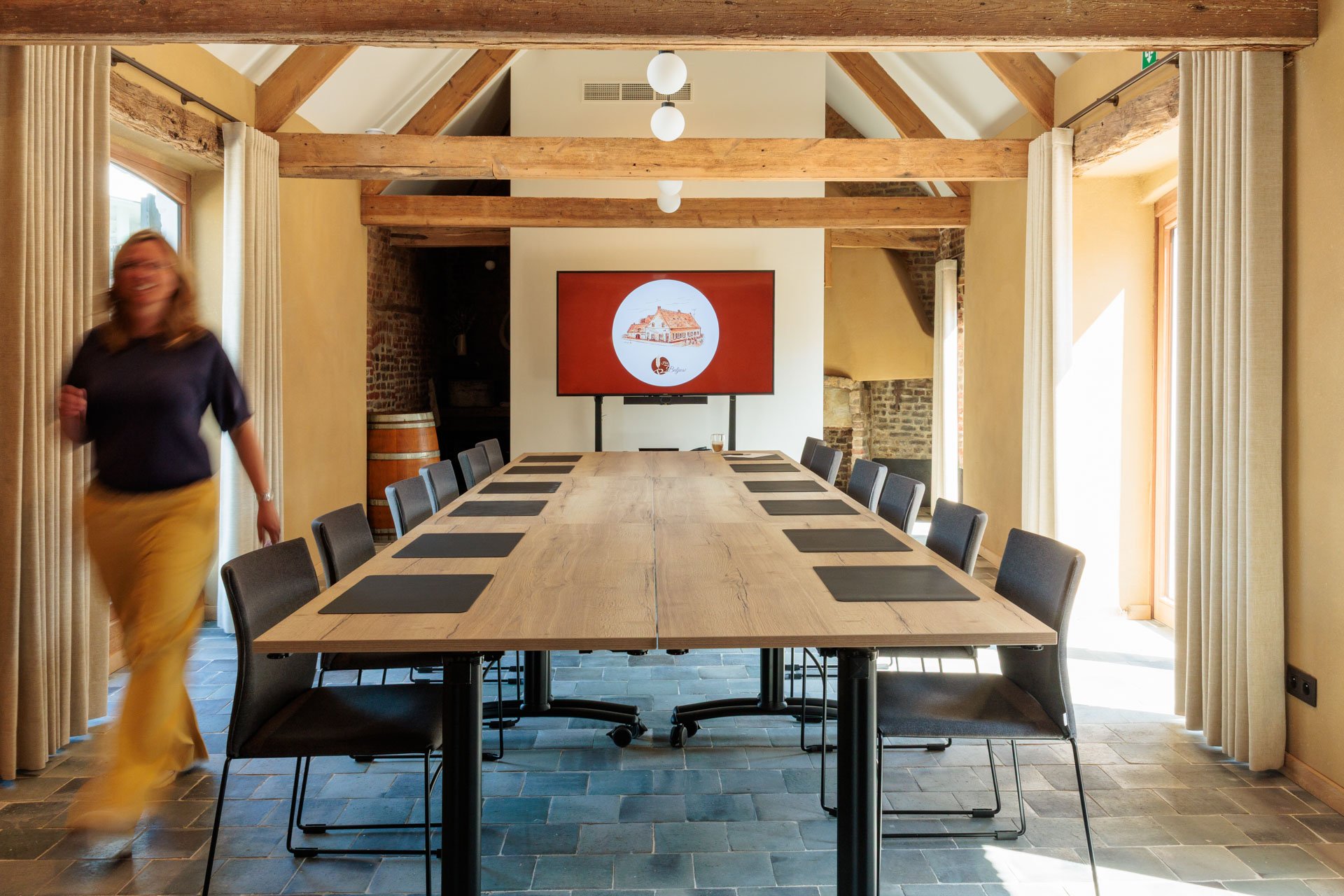 Een vergaderzaal met een grote houten tafel en zwarte stoelen, een scherm met een afbeelding van een traditioneel gebouw, en grote ramen bedekt met beige gordijnen, in een ruimte met houten balken en stenen muren.