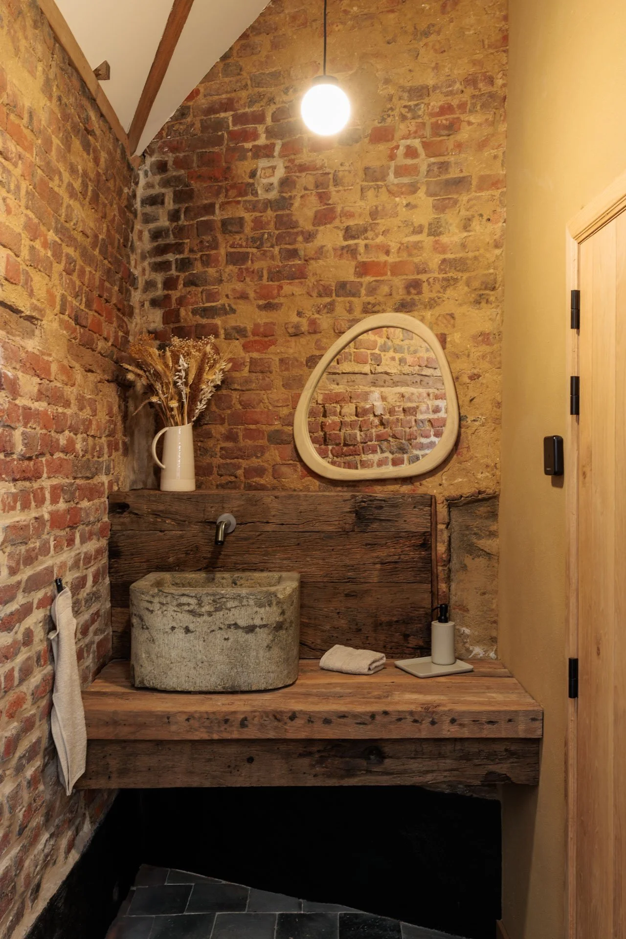 Interieur van een badkamer met een bakstenen muur, een ronde spiegel, een wasbak gemaakt van een oude boomstam, en decoratieve elementen zoals een vaas met gedroogde bloemen. Alles heeft een rustieke en minimalistische stijl.
