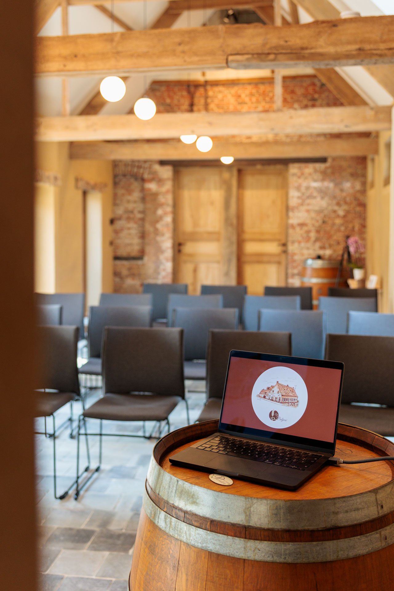 Een vergaderruimte met rijen stoelen, een houten plafond en een laptop op een houten blad. Op de laptop is een logo te zien met een tekening van een huis en de tekst 'De Pelgrim'.
