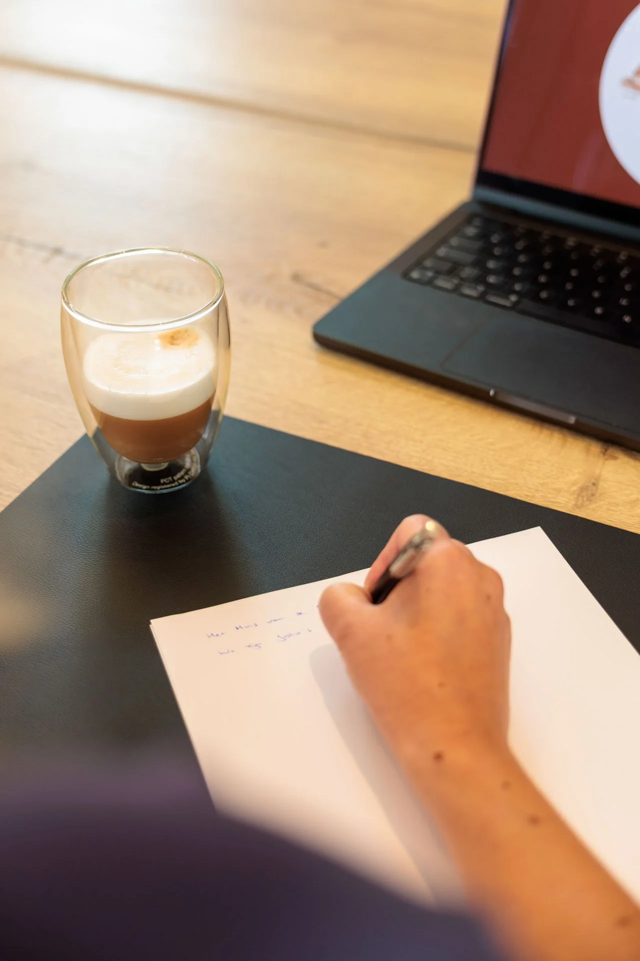 Een hand schrijft op een vel papier, op de tafel ligt een zwart mapje. Op de tafel staat ook een glas met een laag van witte en bruine vloeistof en een open laptop.