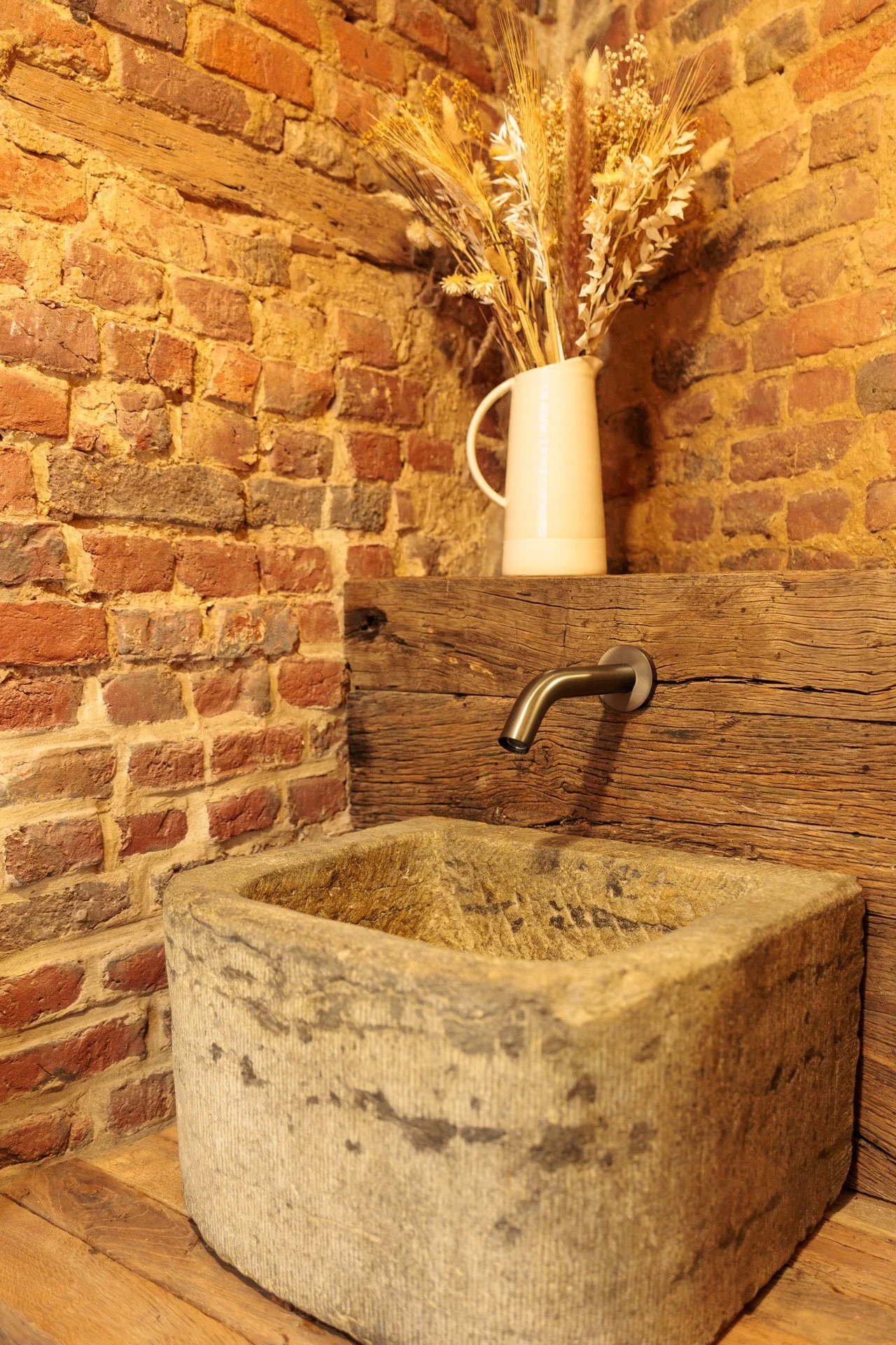 Een rustiek wasbecken van steen met een houten afwerking eromheen, een moderne metalen kraan en een witte vaas met gedroogde bloemen, tegen een bakstenen muur.