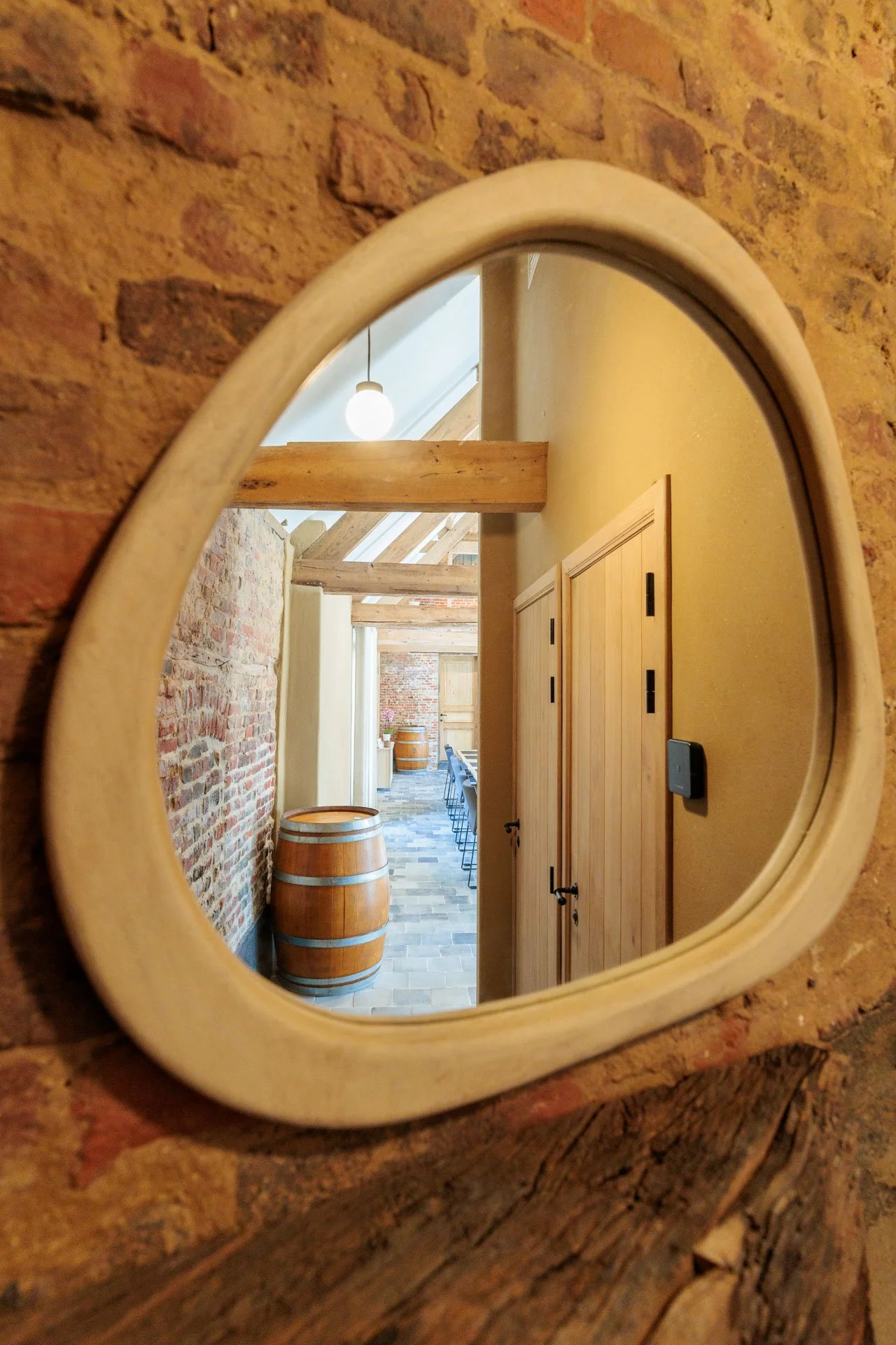Binnenkant van een huis, zichtbaar via een ronde spiegel met een houten rand, waarop een interieur met wijnvaten, houten balken, en een tafel zichtbaar is.