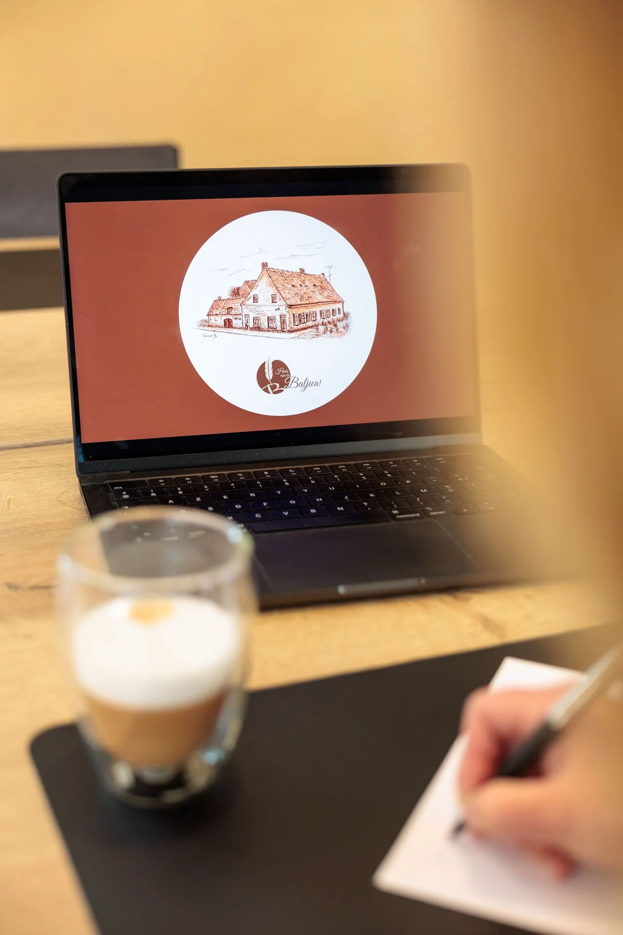 Een laptop op een houten tafel met een logo van een huis in een cirkel op het scherm, een glas koffie op een zwarte onderlegger en iemand die schrijft op een stuk papier.