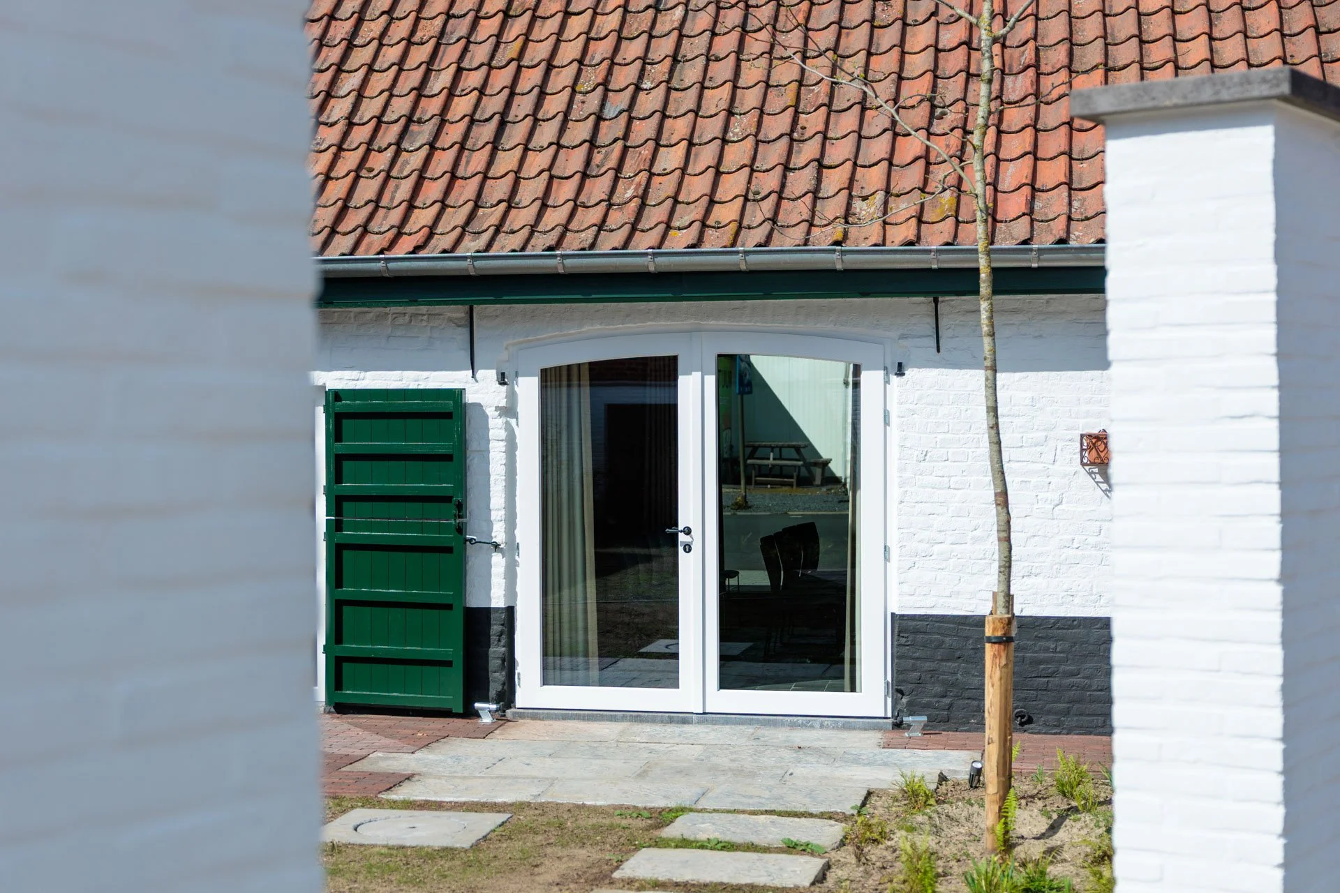 De voorkant van een witte woning met een groene deur en een glazen deur die leidt naar binnen, met een rode dakpan gedekt dak en een jonge boom geplant naast de deur.