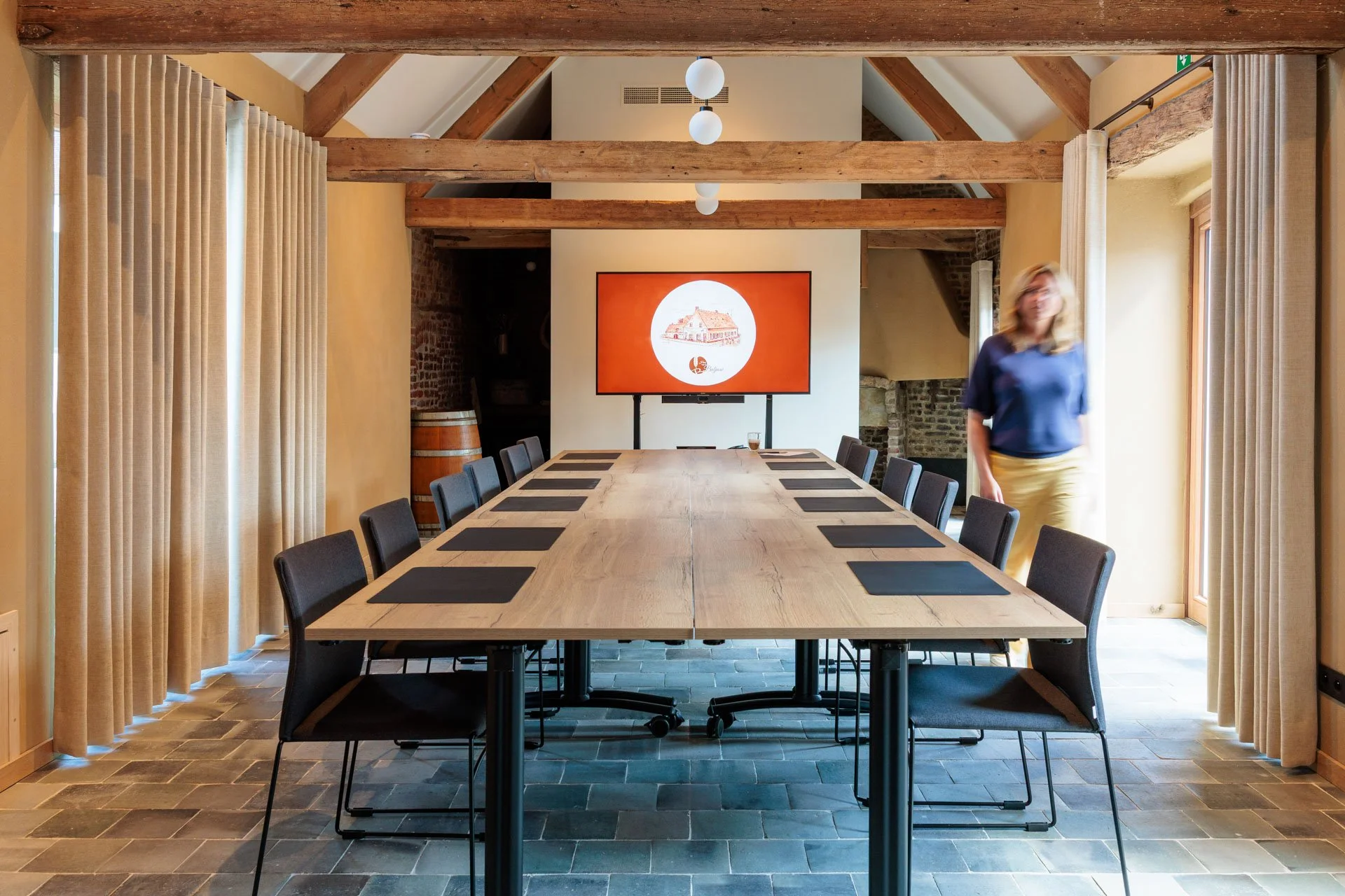 Een vergadertafel in een vergaderruimte met 14 zwarte stoelen, een grote tv-scherm aan de muur, houten balken en gordijnen aan de zijkanten.