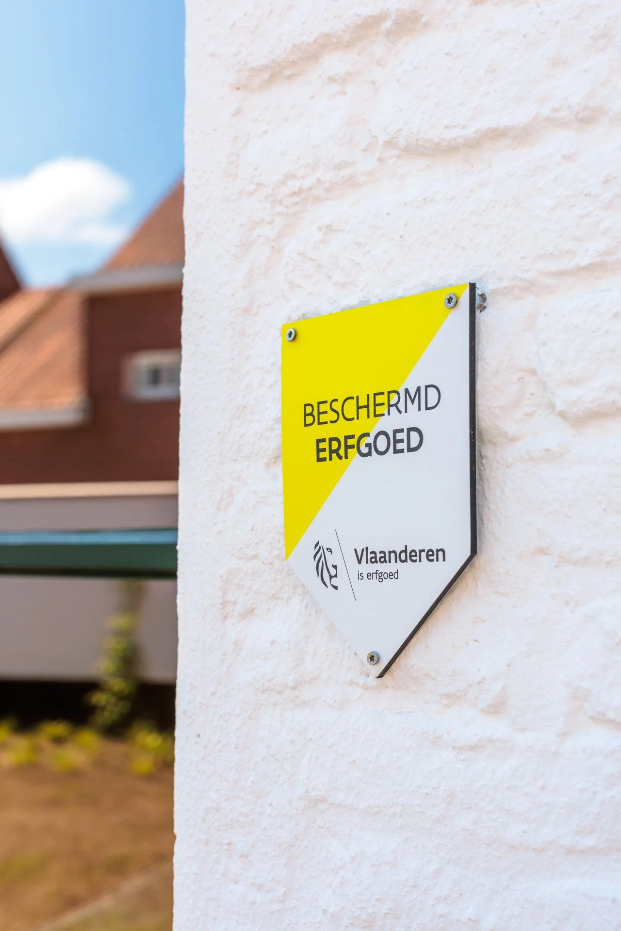 Een waarschuwingsbord aan een witte muur met de tekst "BESCHERMD ERFGOED" en het logo van Vlaanderen, met een achtergrond van een huizencomplex en een blauwe lucht.