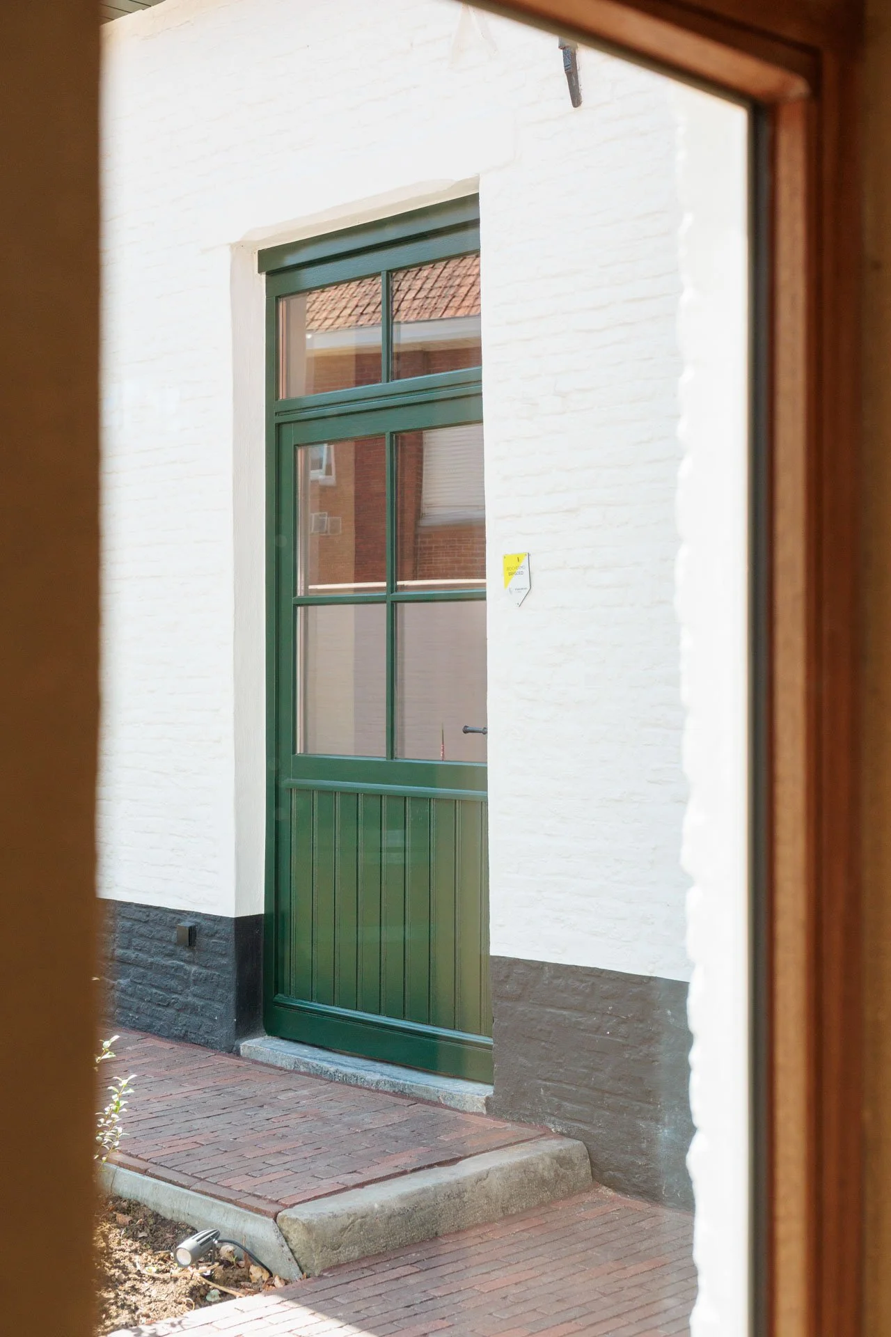 De afbeelding toont een groene deuropening met een groen raam boven en onder, gezien vanaf binnen door een raam. Buiten is een witte muur met een klein struikje en een verlichte buitenlamp op de grond.