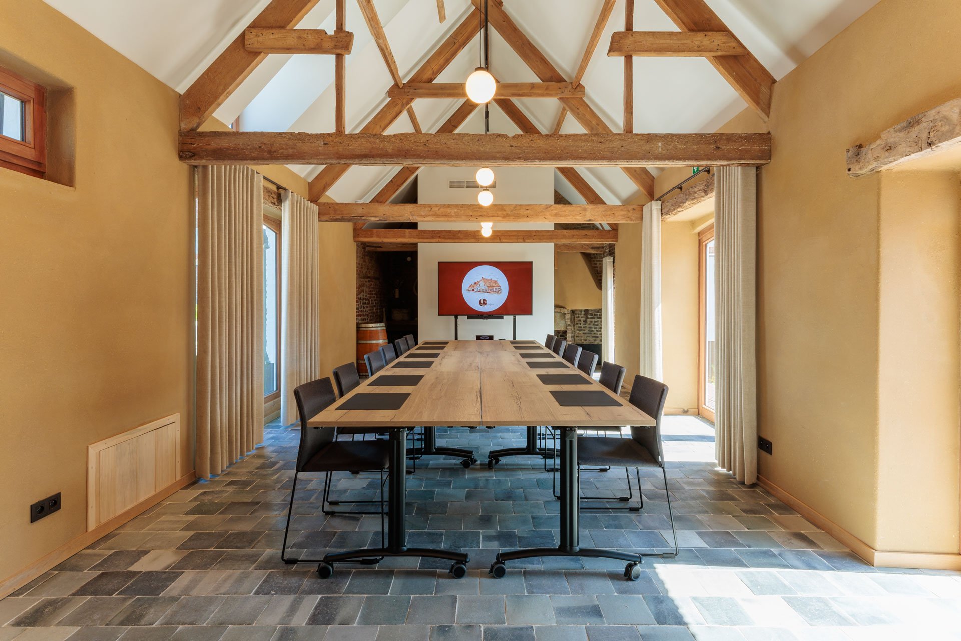 Een vergaderruimte met een lange houten tafel, omgeven door zwartige bureaustoelen, en een muur met een scherm, verwarmingsradiator, grote ramen en houten balken in het plafond.