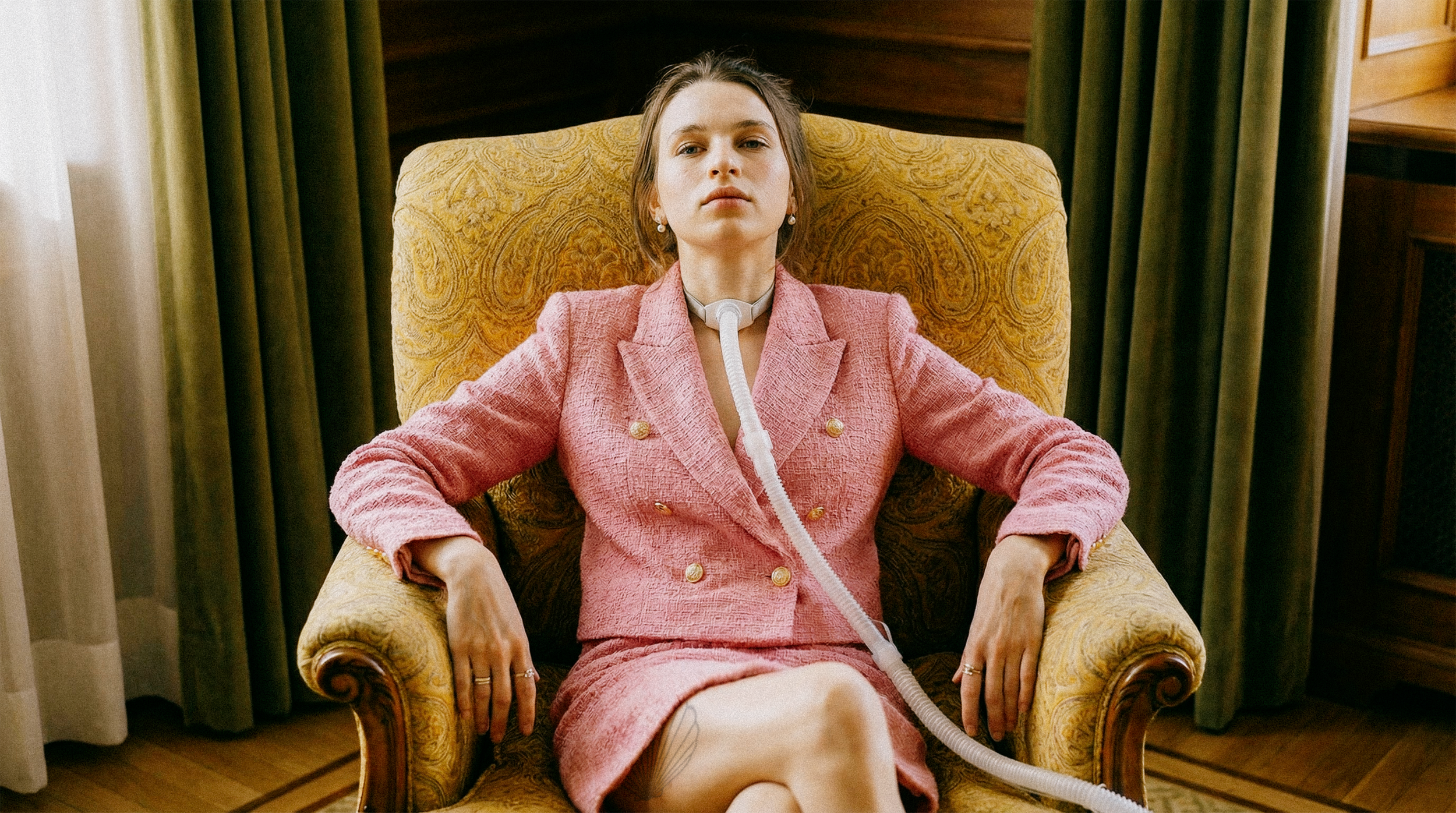 A woman wearing a pink suit sitting in a yellow armchair with medical tubing connected to her neck, in a room with green curtains and wooden walls.