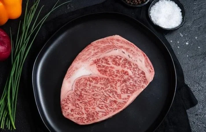 Raw marbled beef steak on a black plate with salt, green onions, and bell peppers in the background