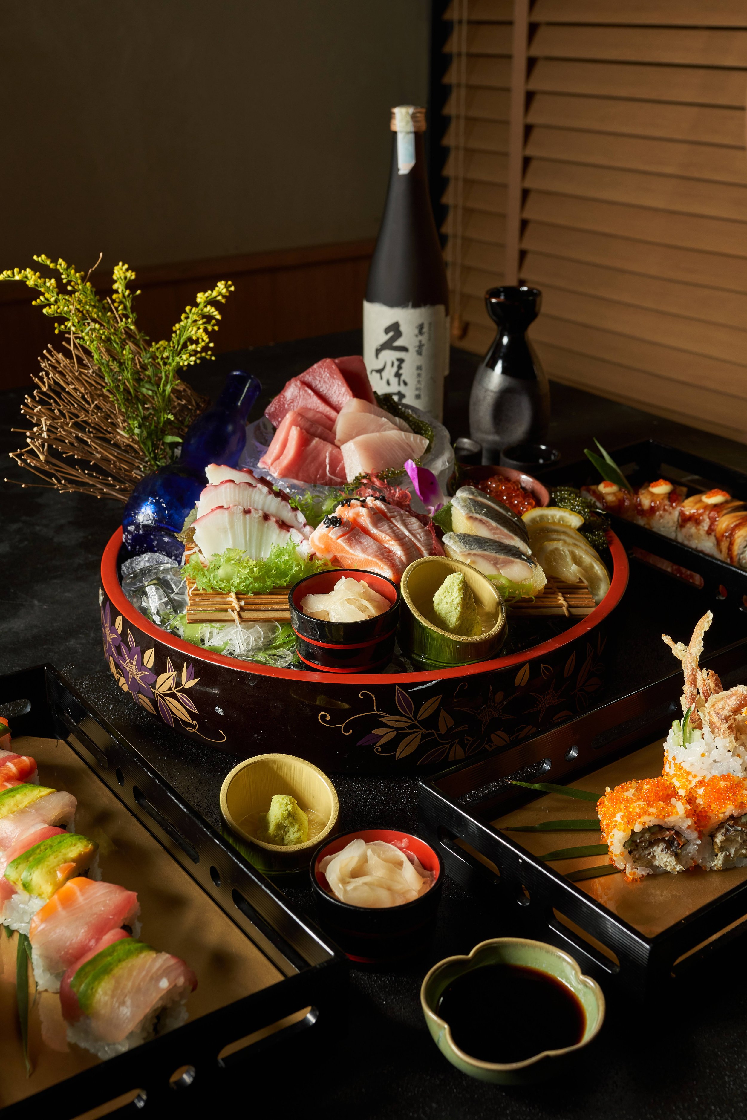 A variety of sashimi and sushi arranged on a tray with garnishes, wasabi, and soy sauce, accompanied by a bottle of sake and a sake bottle on a dark table.