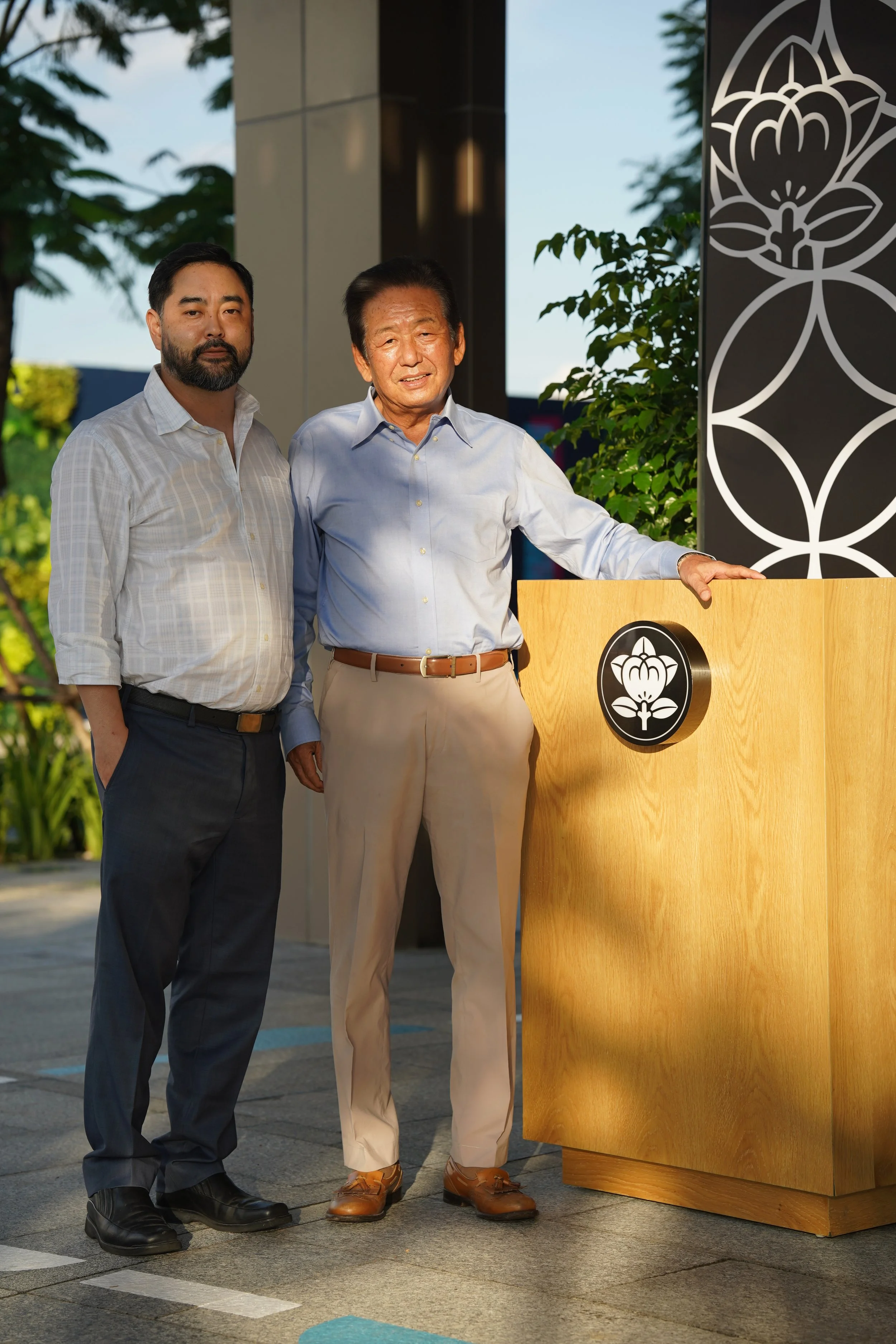 Two men standing outdoors near a podium featuring a lotus flower symbol, with trees and a building in the background. One man has a beard and wears a white shirt, and the other is older, dressed in a light blue shirt and tan pants.